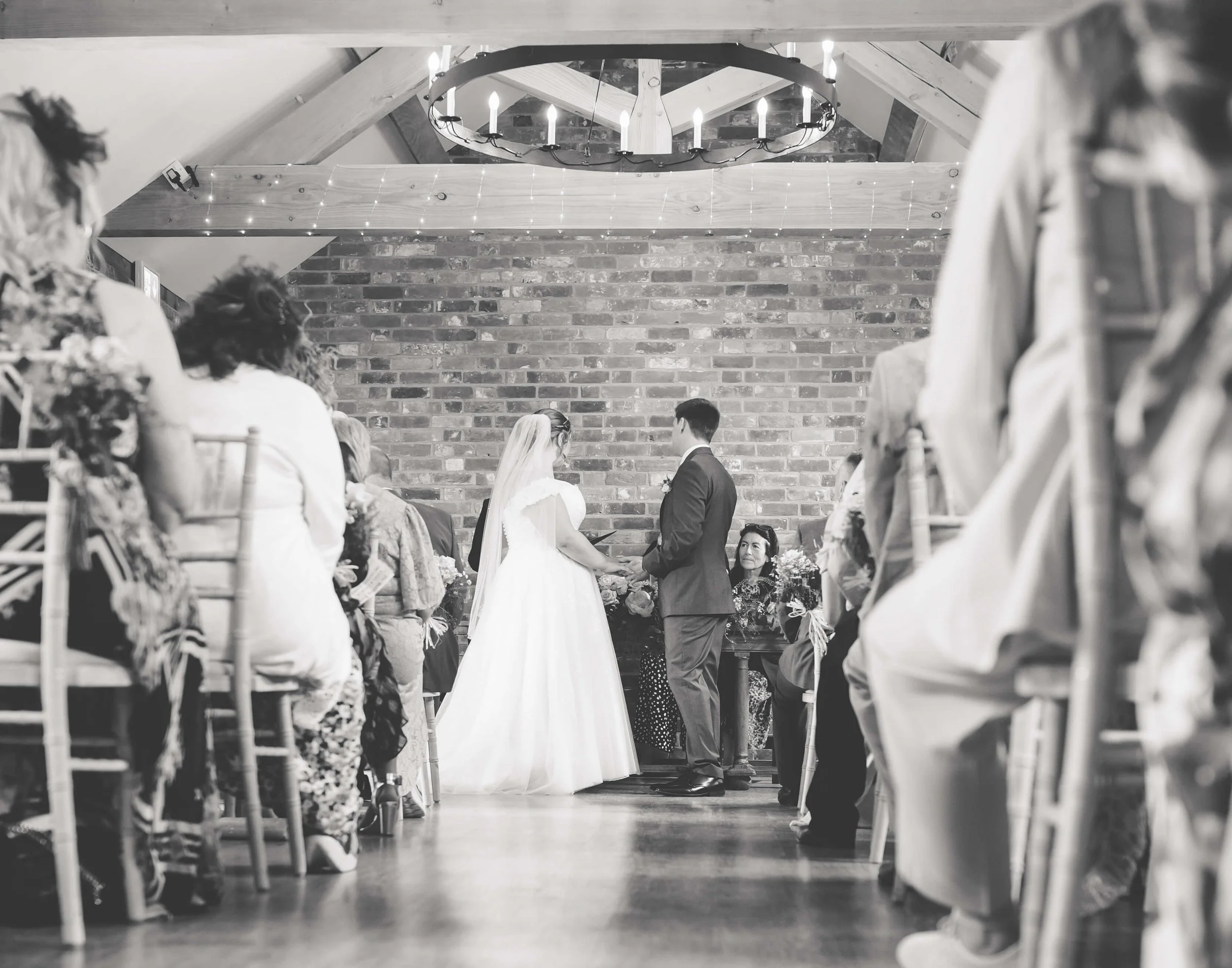 A black-and-white photo of a wedding ceremony with the bride and groom holding hands, facing each other, in front of a brick wall, with guests seated on either side and a woman sitting behind a table.