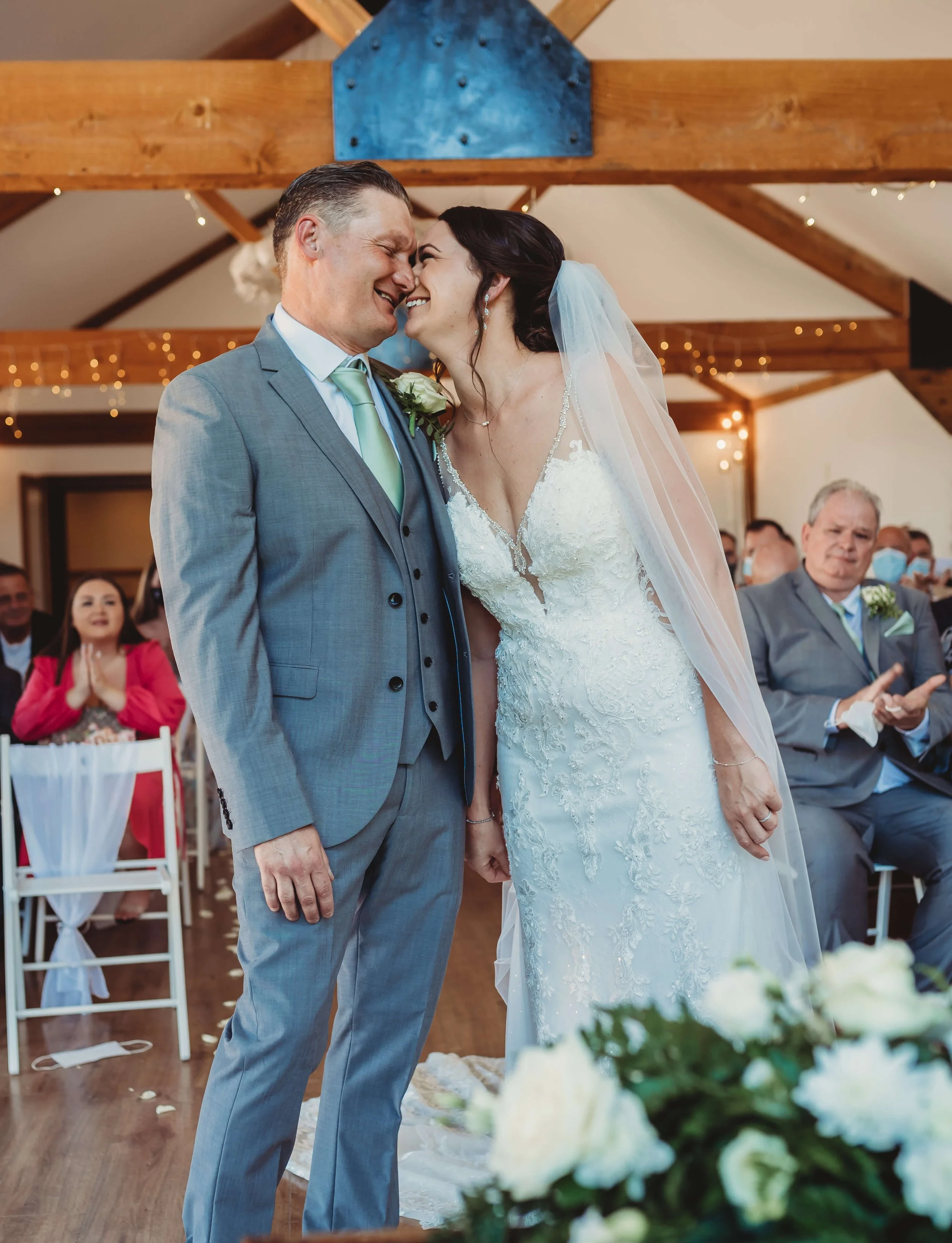 Bride and groom sharing a kiss during their wedding ceremony in a rustic indoor venue, with guests watching and smiling in the background.