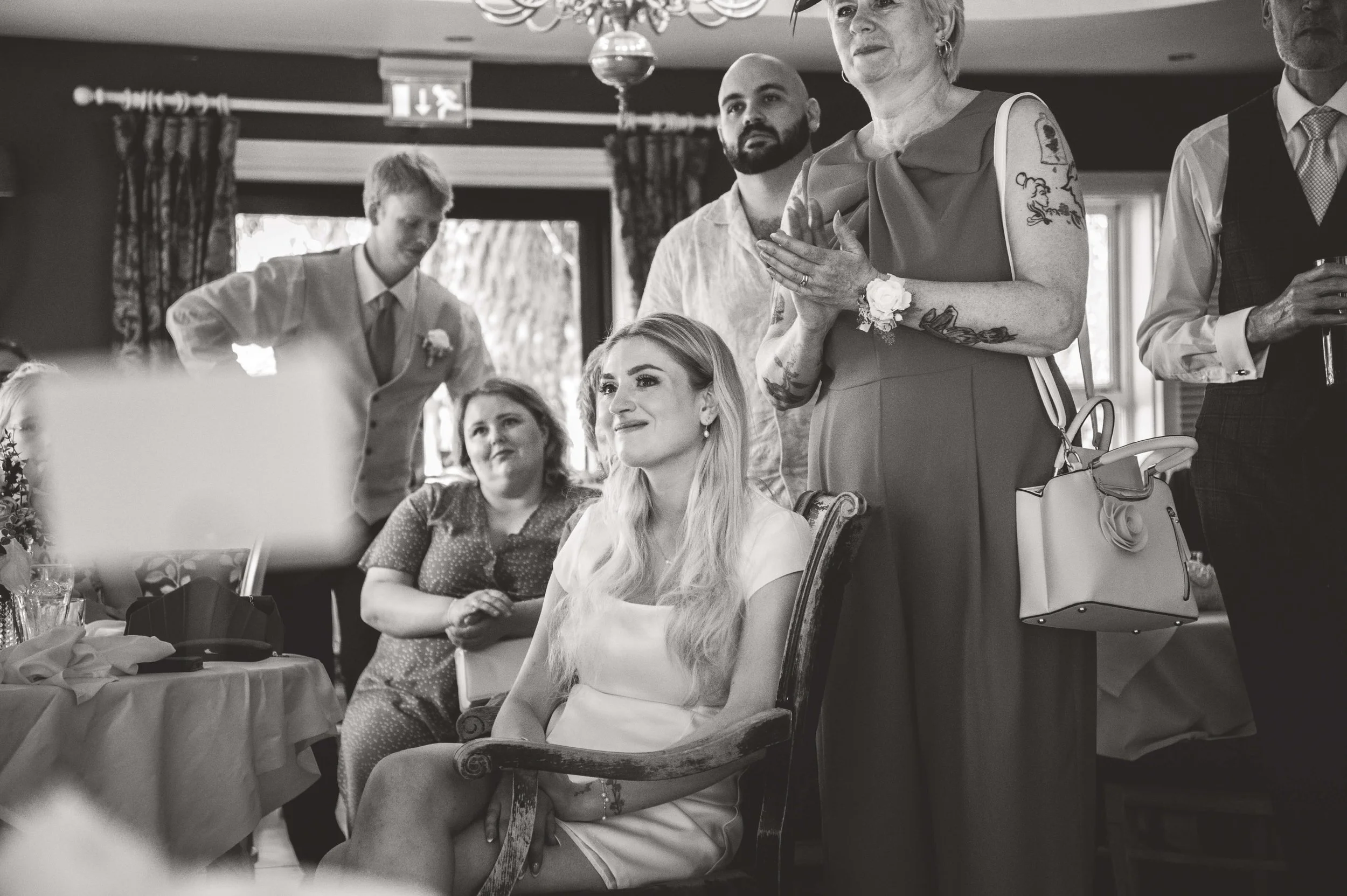 A black and white photo of a woman sitting on a wooden chair during a celebration or event. Several people are standing around her, some with hands on their chest, inside a decorated room with curtains and a chandelier.