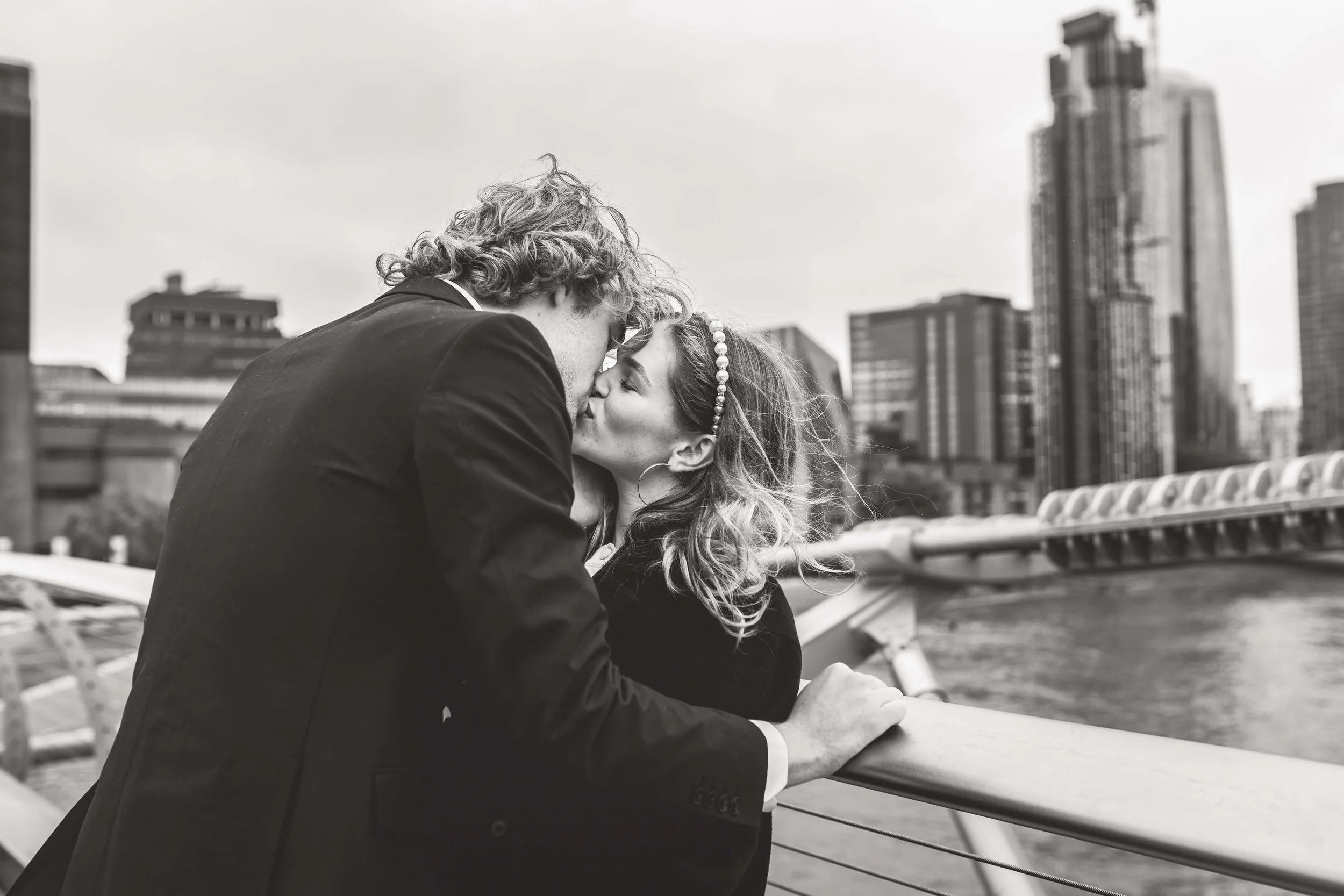 A couple kissing on a city bridge, black and white photo with tall buildings in the background.