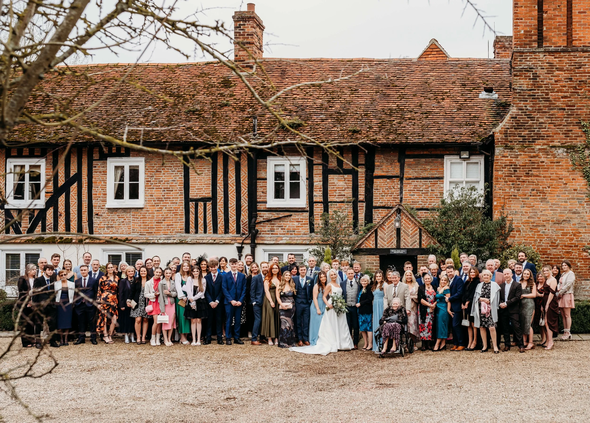 A large group of wedding guests gathered outside a historic brick building with a red tiled roof, black timber framing, and white windows, gathered for a group photo.