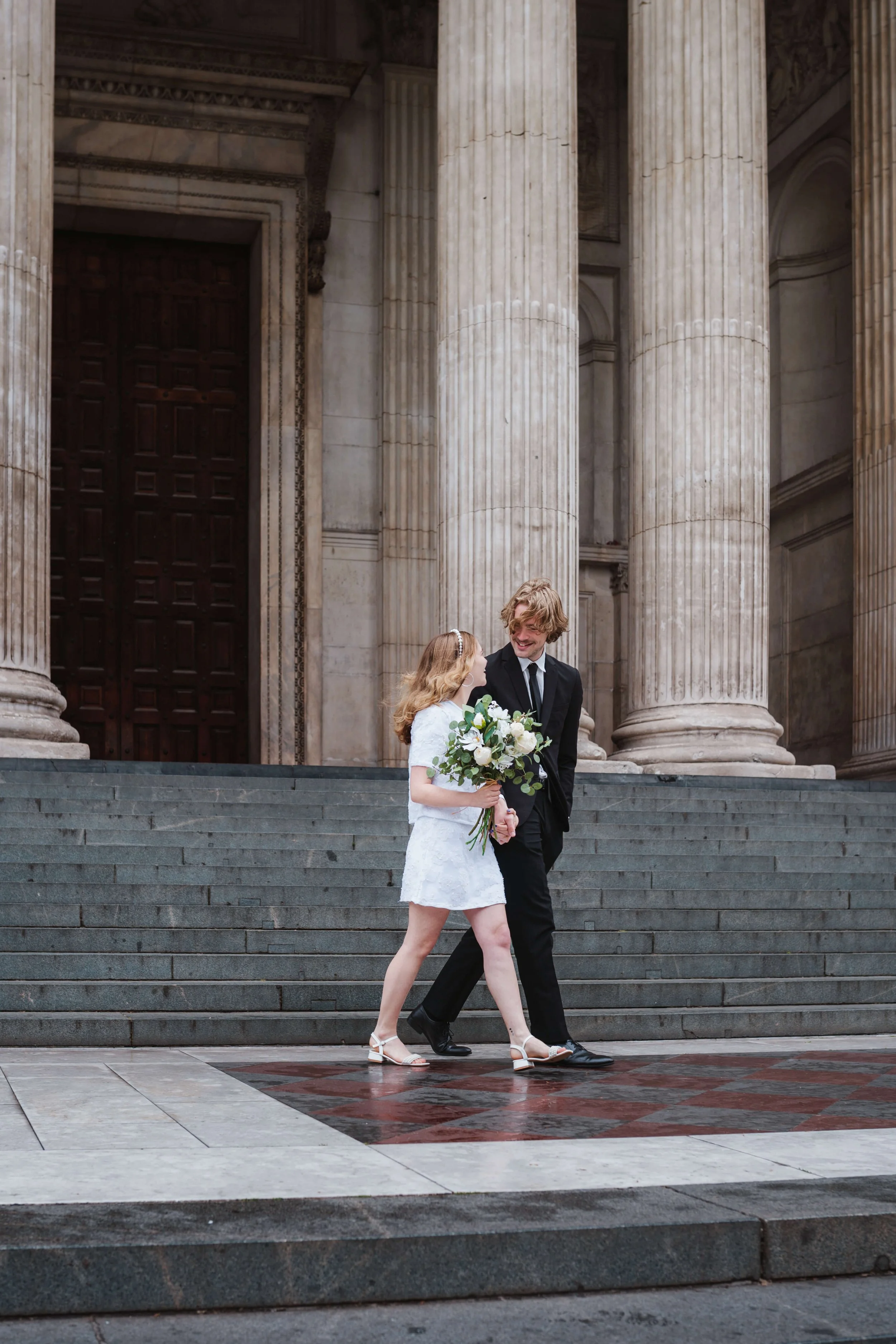 A young couple, dressed in wedding attire, walking hand in hand down steps outside a grand neoclassical building with large columns, holding a bouquet of flowers.