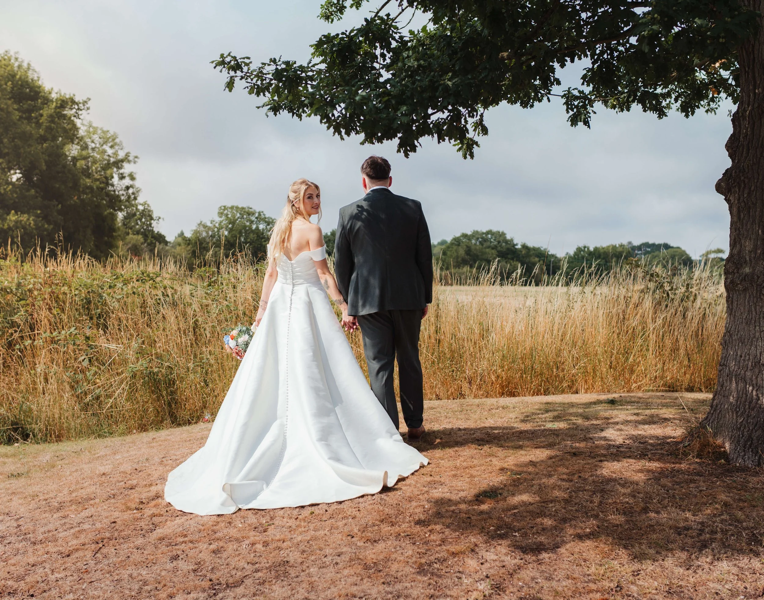 A bride in a white wedding gown holding a bouquet and a groom in a black suit holding hands, walking under a large tree in an outdoor field.