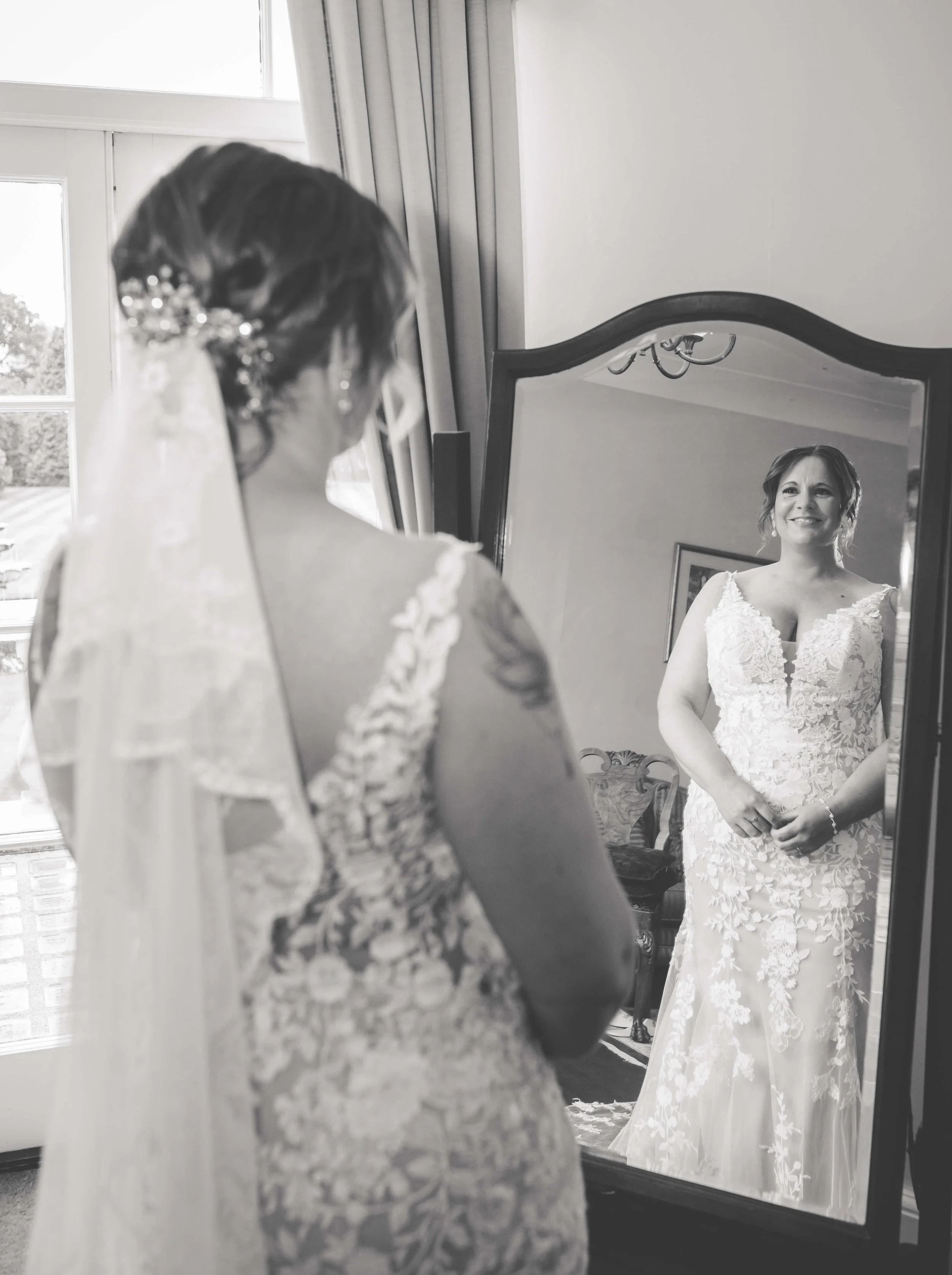 A bride looking at herself in a mirror, smiling, wearing a lace wedding gown with floral embroidery and a veil, in a room with a window and curtains.