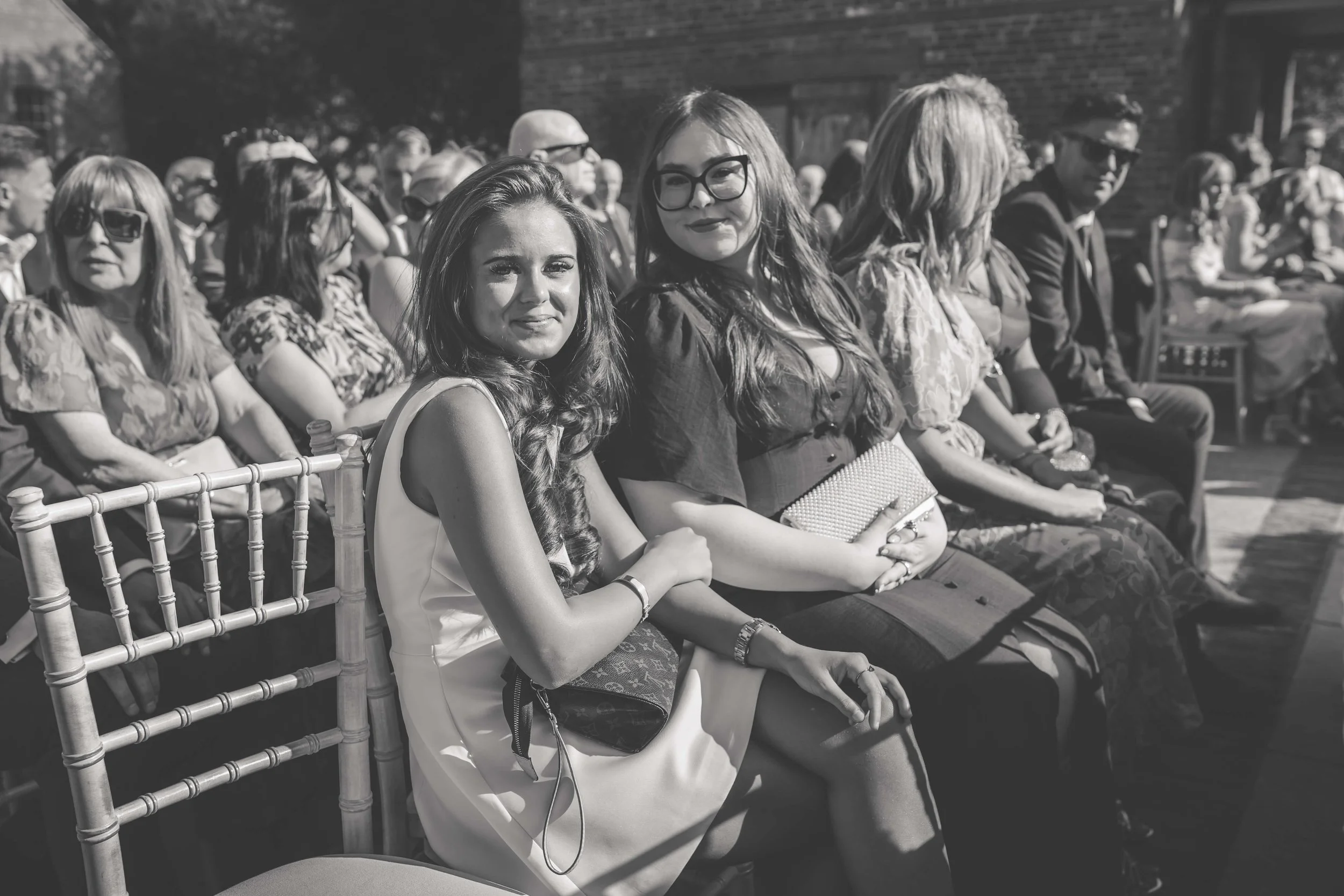 Black and white photo of a group of people sitting outdoors, with two young women in the foreground smiling at the camera. The woman on the left has long, wavy hair, wearing a sleeveless dress, and the woman on the right has long hair, glasses, and i