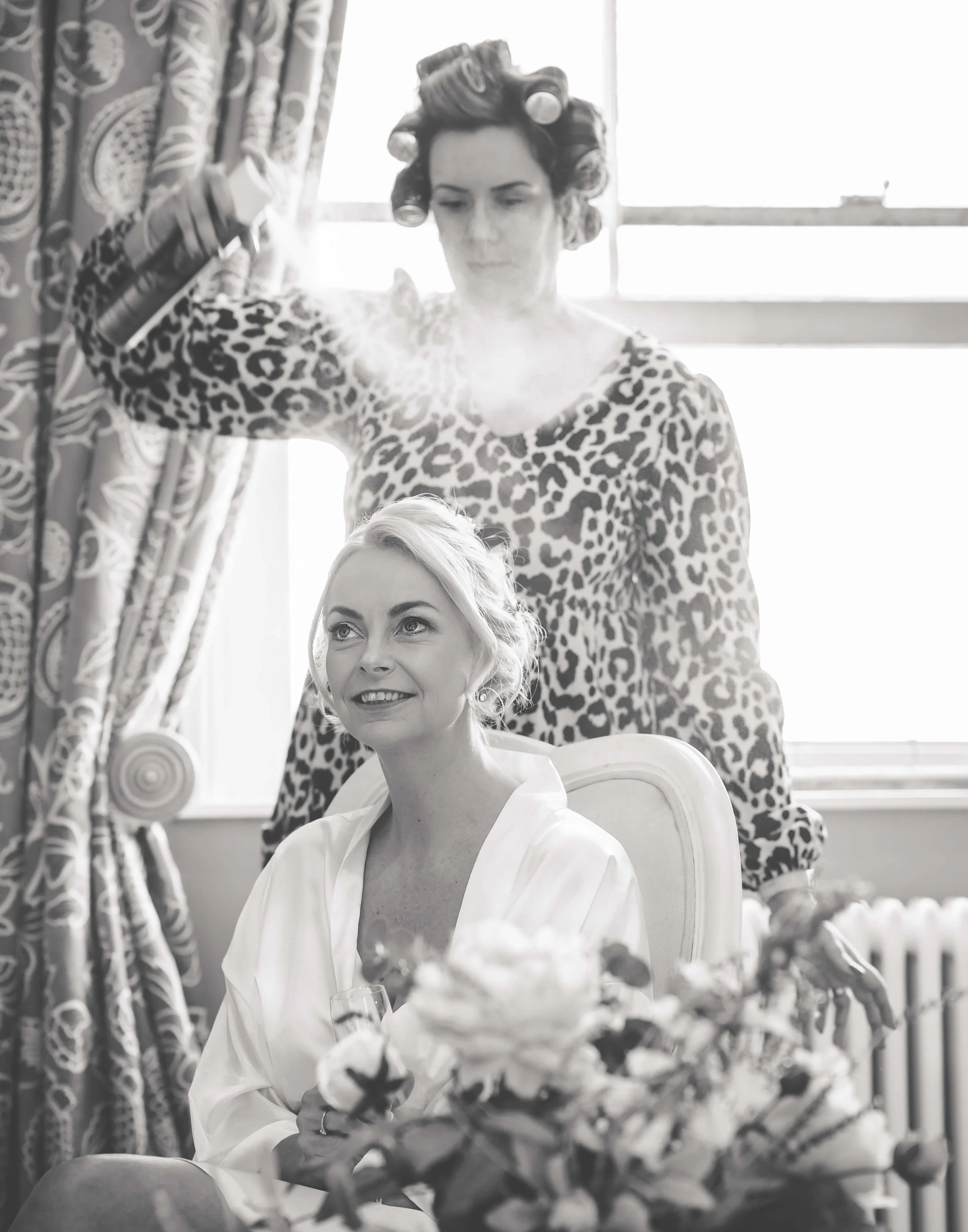 A woman with short blonde hair, wearing a white robe, is sitting in a chair holding a flower, while another woman with dark hair and hair rollers, wearing a leopard print top, is using a spray to style her hair in a bright room with patterned curtain
