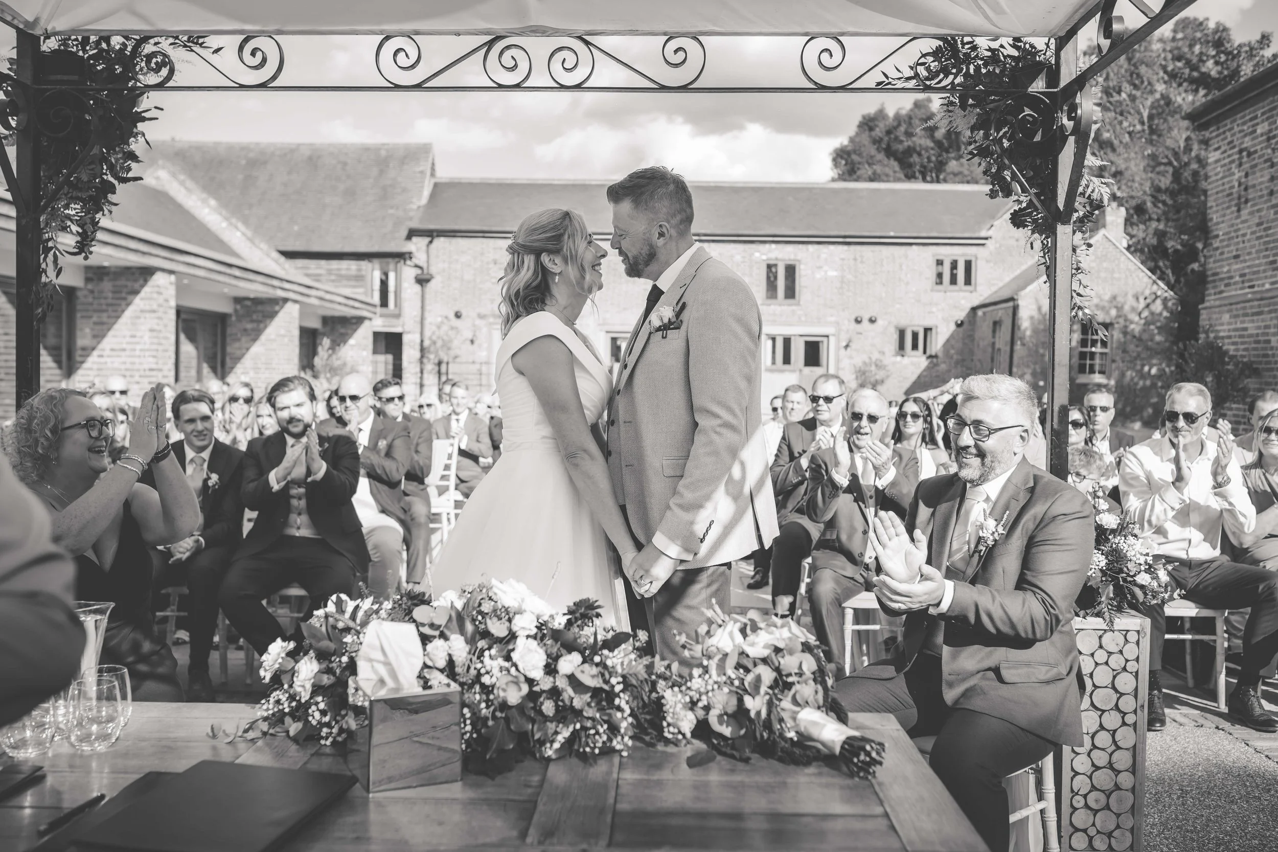 A black-and-white photo of a wedding ceremony outdoors with a bride and groom holding hands and facing each other under a decorative arch, surrounded by seated and clapping guests.