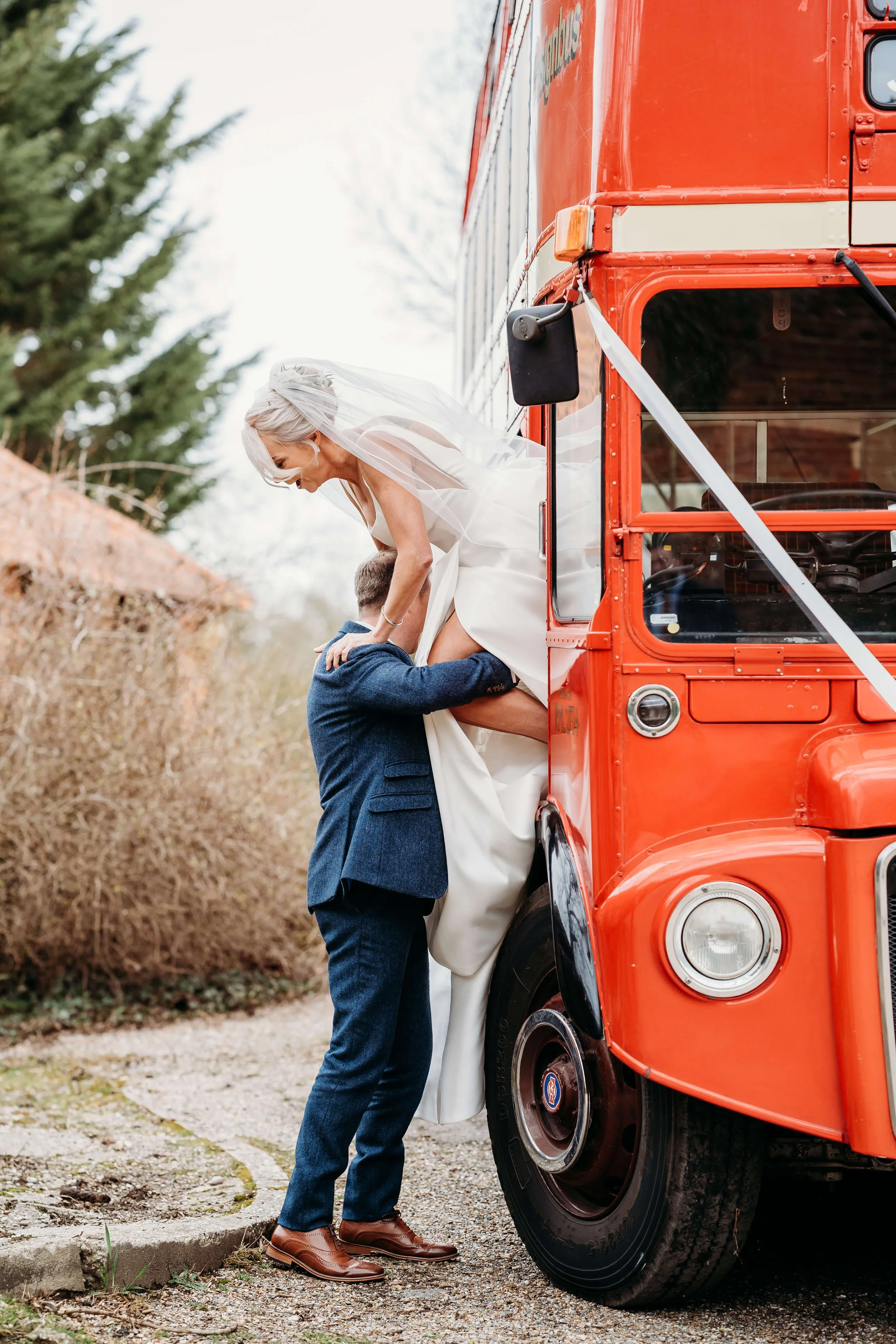 A wedding couple is having their wedding photos taken outdoors next to a vintage orange bus. The groom is lifting the bride, who is dressed in a white wedding gown and veil, while the groom wears a dark suit. The scene is on a gravel path surrounded 