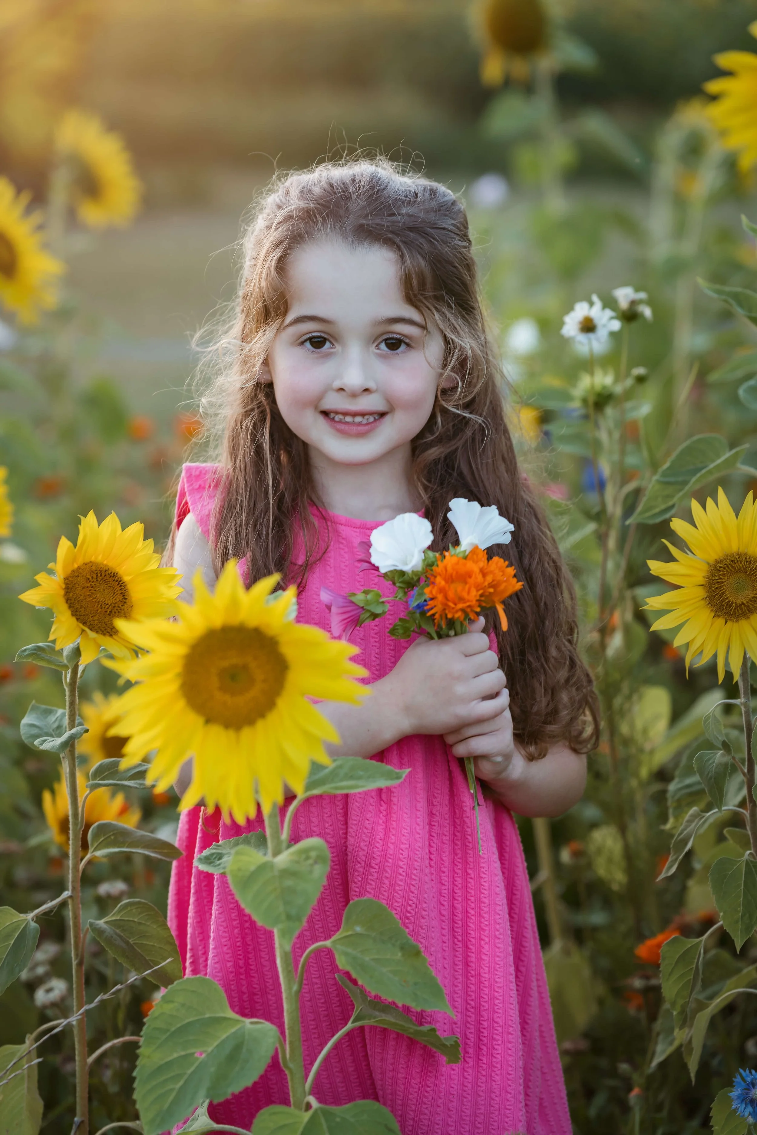 A young girl with long, curly brown hair, wearing a pink dress, standing in a sunflower field, holding a small bouquet of white, orange, and pink flowers, smiling at the camera during sunset.