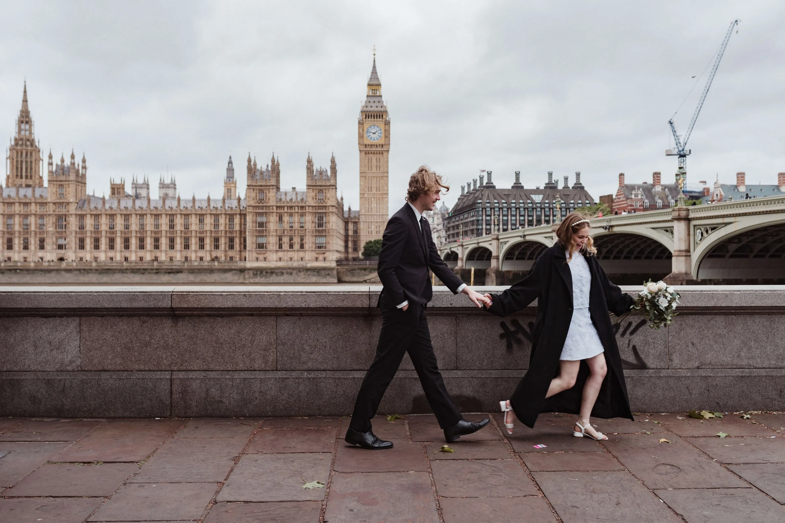 A couple walking hand in hand along a bridge with the Palace of Westminster and Big Ben in London in the background. The woman is holding a bouquet of flowers and wearing a white dress with a black coat, and the man is wearing a black suit.