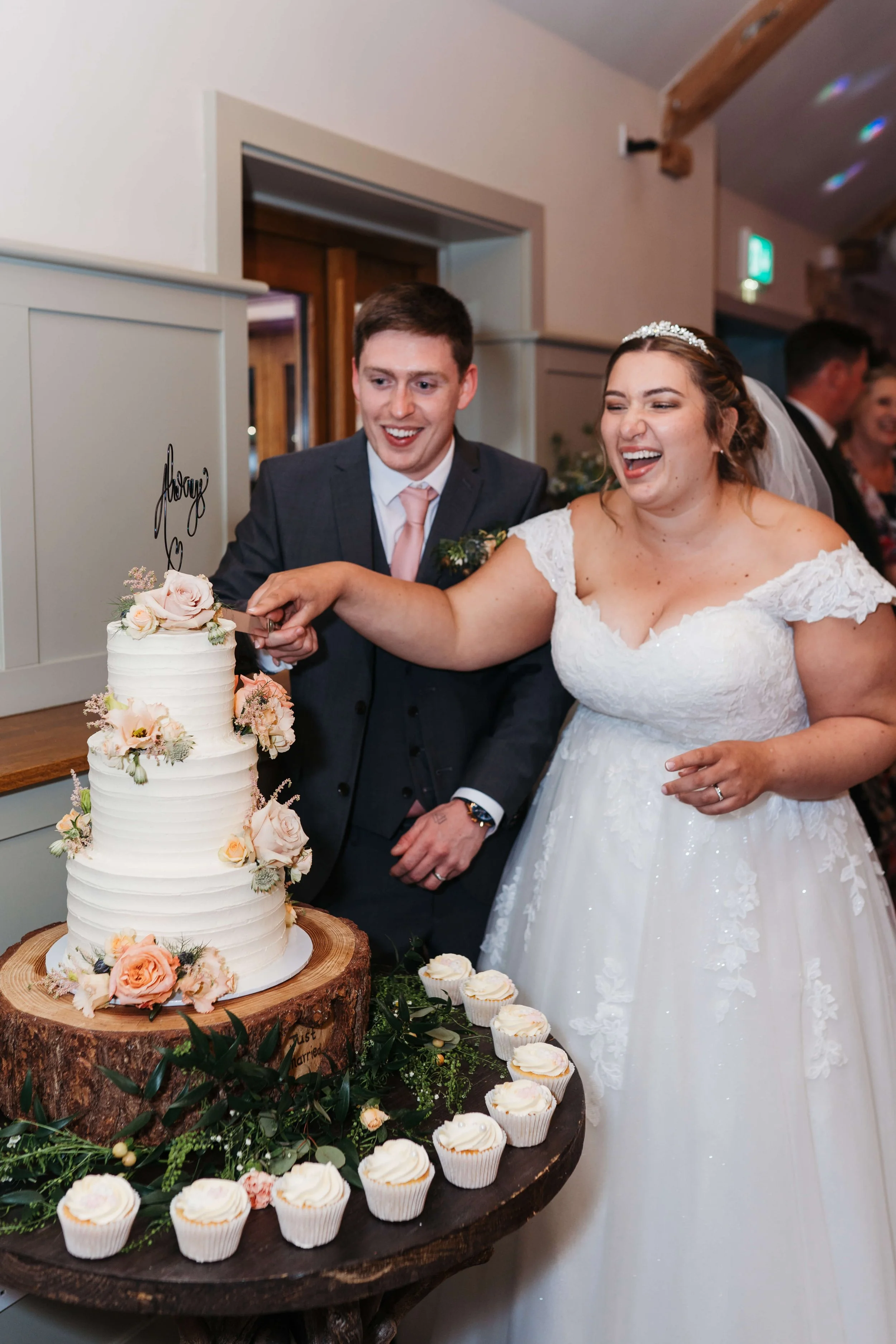 Bride and groom cutting their wedding cake together at the reception, surrounded by cupcakes and floral decorations.