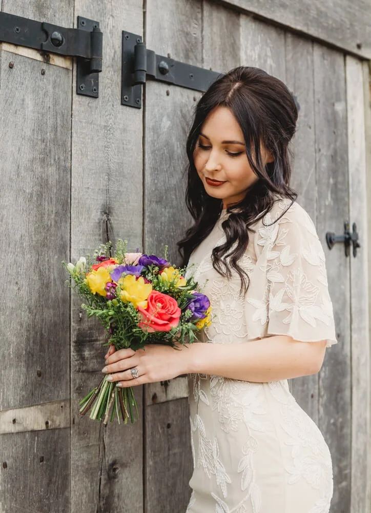 A woman with dark, styled hair holding a colorful bouquet of flowers in front of a wooden door.
