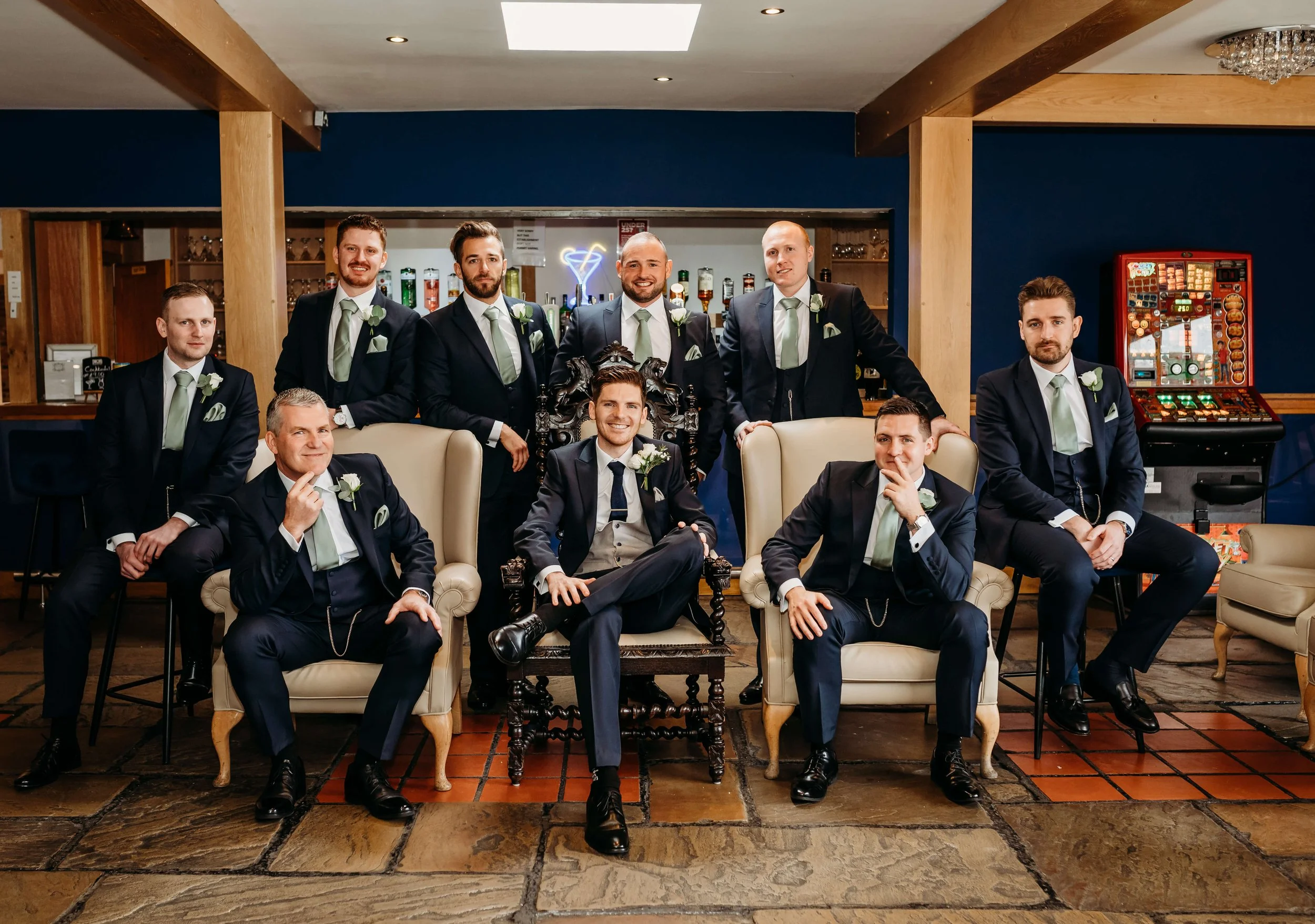 Group of eleven men dressed in formal suits, posing for a photo in a bar, with a background of drink bottles and a gaming machine.