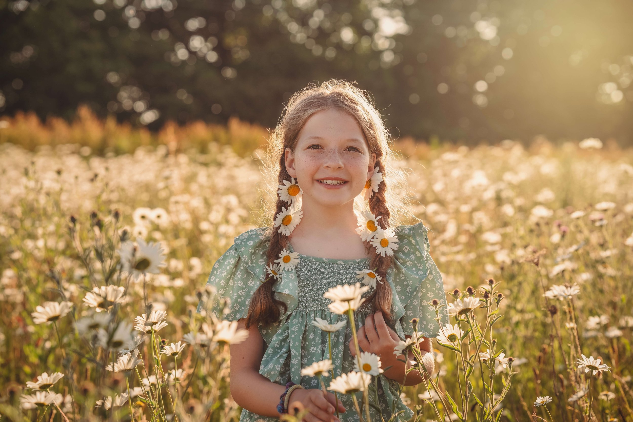 A young girl with red hair in pigtails decorated with daisies, smiling and standing in a field of daisies during golden hour.
