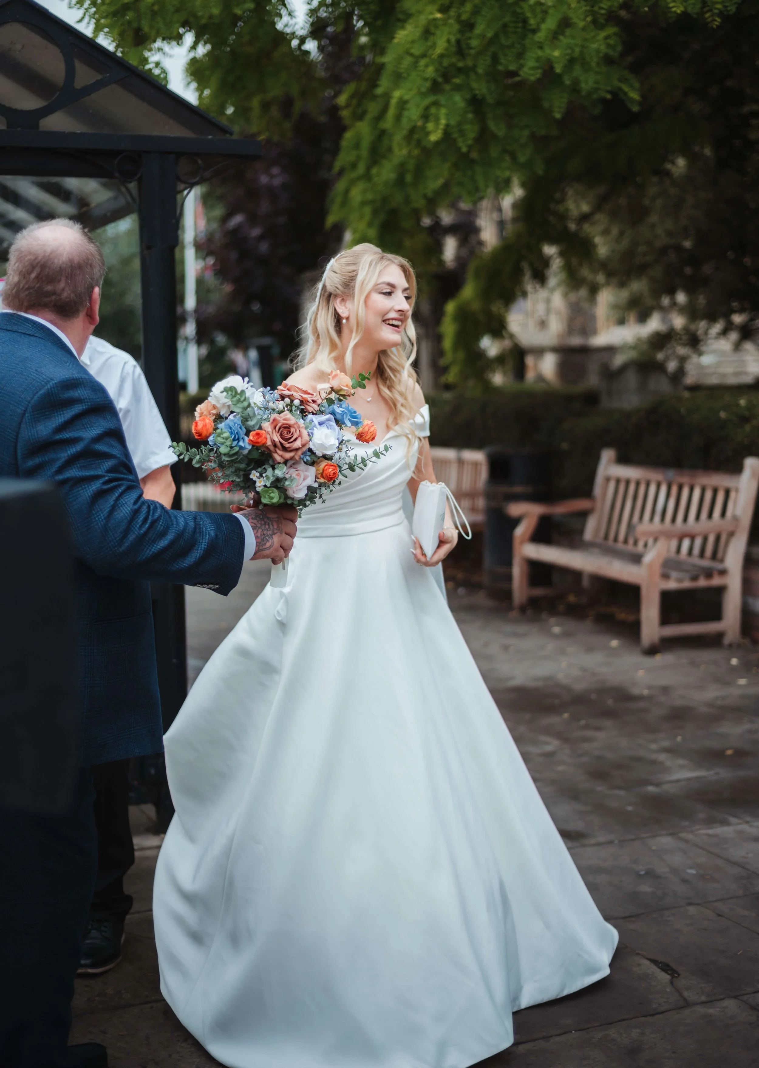 A bride in a white wedding gown holding a bouquet of colorful flowers, smiling and walking outdoors with two men, during a wedding ceremony or celebration.