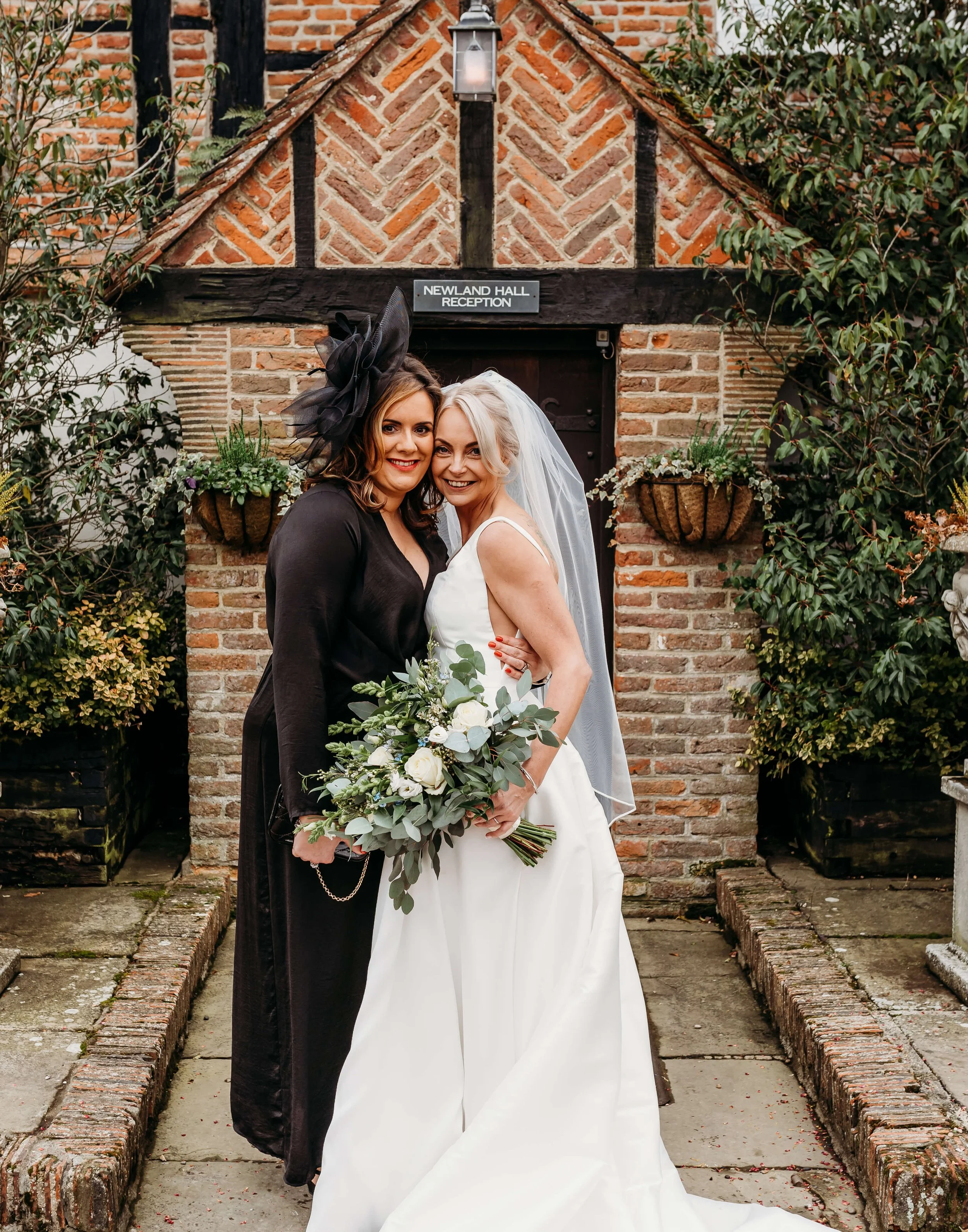 Two women, one in a white wedding dress holding a bouquet of white roses and greenery, and the other in a black dress with a large black fascinator, standing in front of a brick building with a sign that reads 'Newland Hall Reception'.
