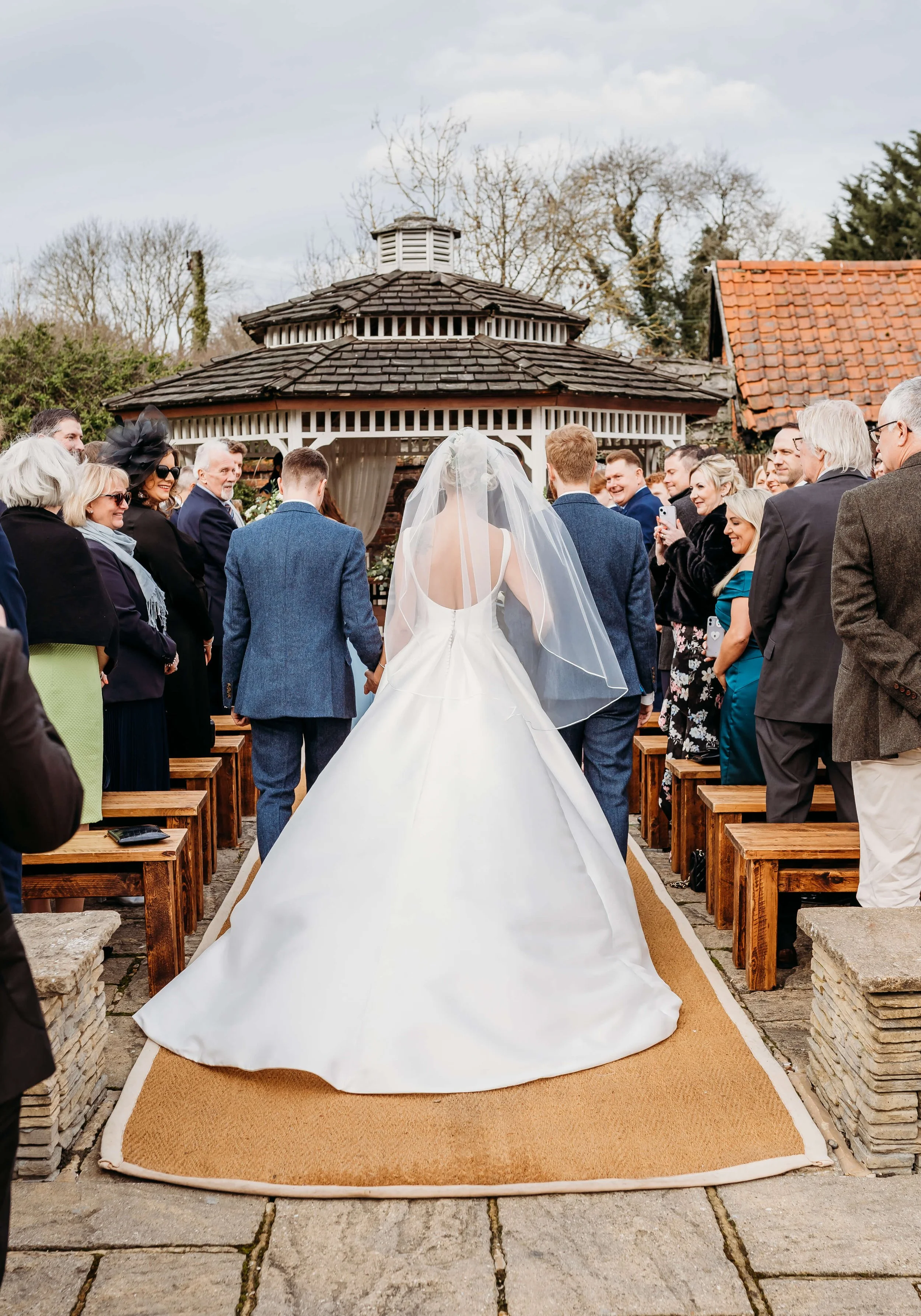 Wedding ceremony with bride in white gown and veil walking down aisle with guests on either side, outdoor setting with gazebo in background.