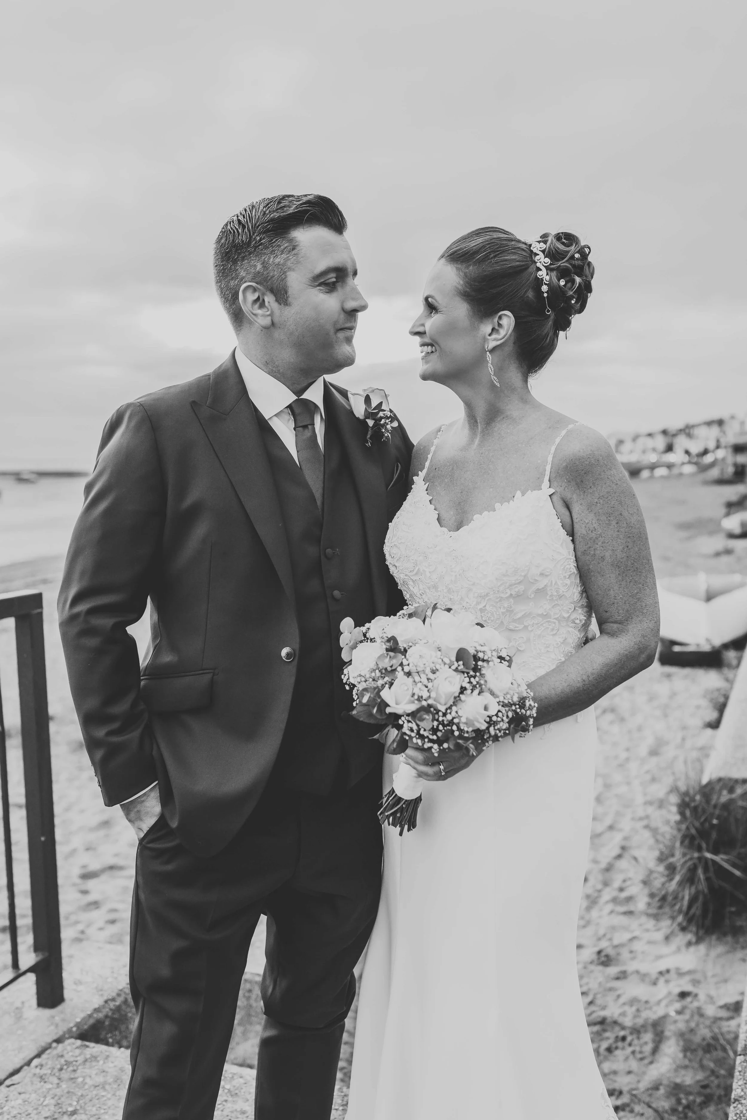 Black and white photo of a bride and groom at the beach, facing each other and smiling, with the ocean and cloudy sky in the background.