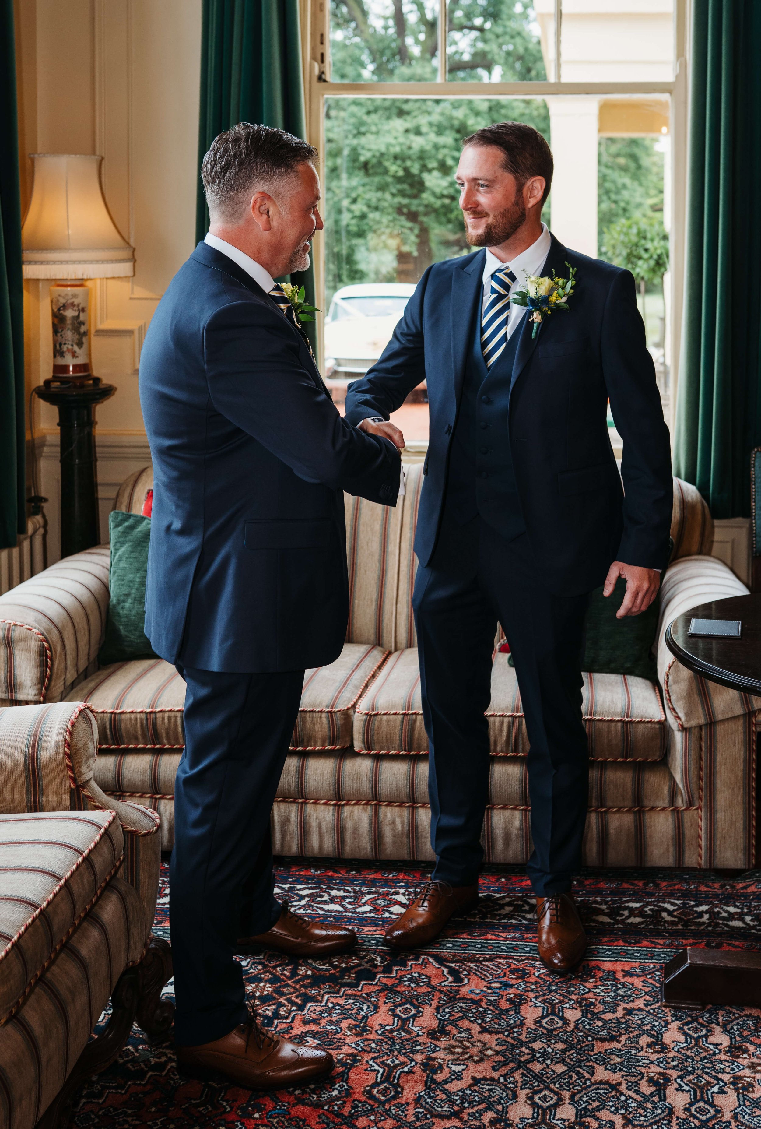 Two men in dark suits shaking hands in a room with green curtains, a striped sofa, and a patterned rug, with a large window showing trees outside.