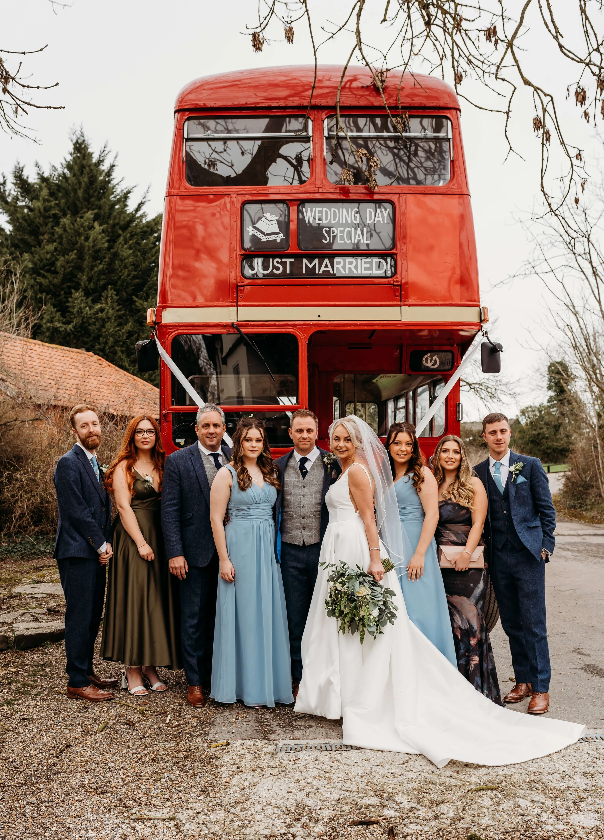 A wedding group photo with the bride and groom in front of a red double-decker bus, which has signs saying "Wedding Day Special" and "Just Married." The bride is wearing a white wedding dress and holding a bouquet of greenery, and the groom is dresse