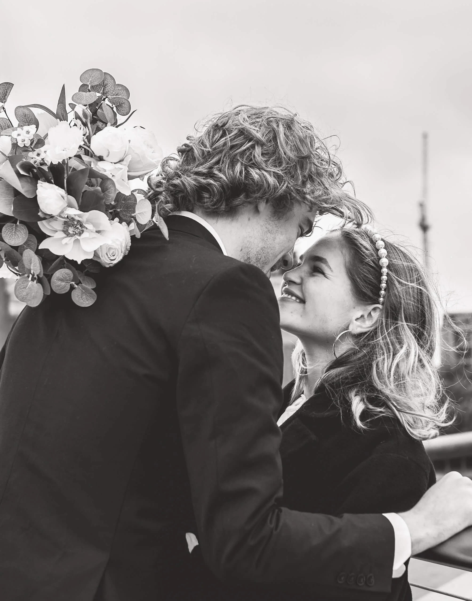 Black and white photo of a couple close together, smiling and about to kiss, with the woman wearing a headband and hoops and the man holding a bouquet of flowers over his shoulder.