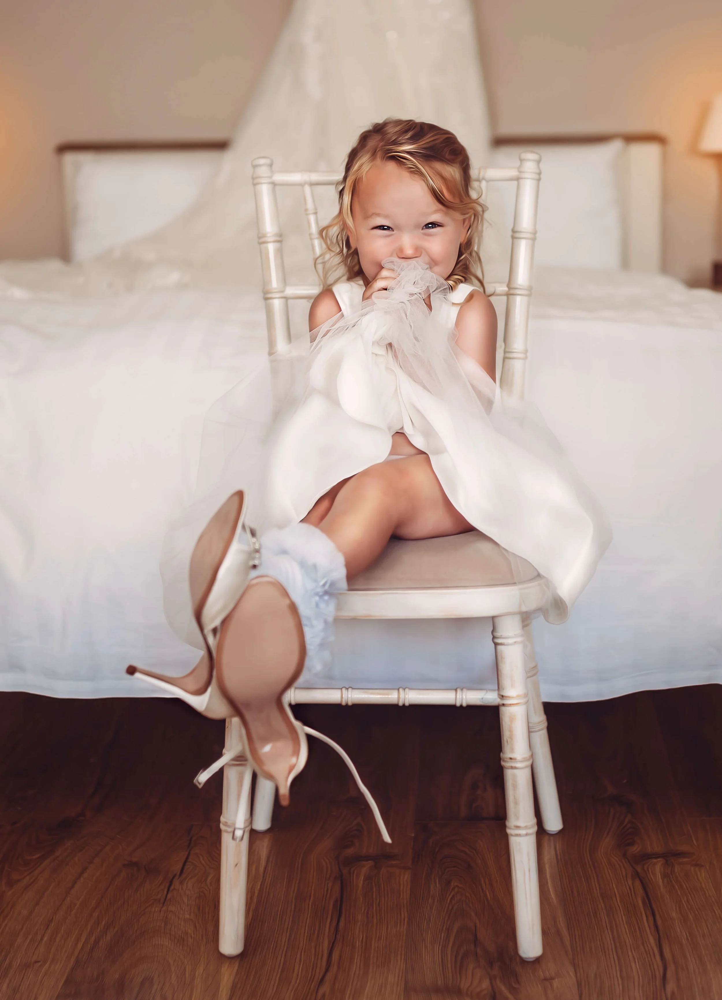 A young girl sitting on a chair in front of a bed, wearing a white dress and high heels, smiling and holding the dress near her face.