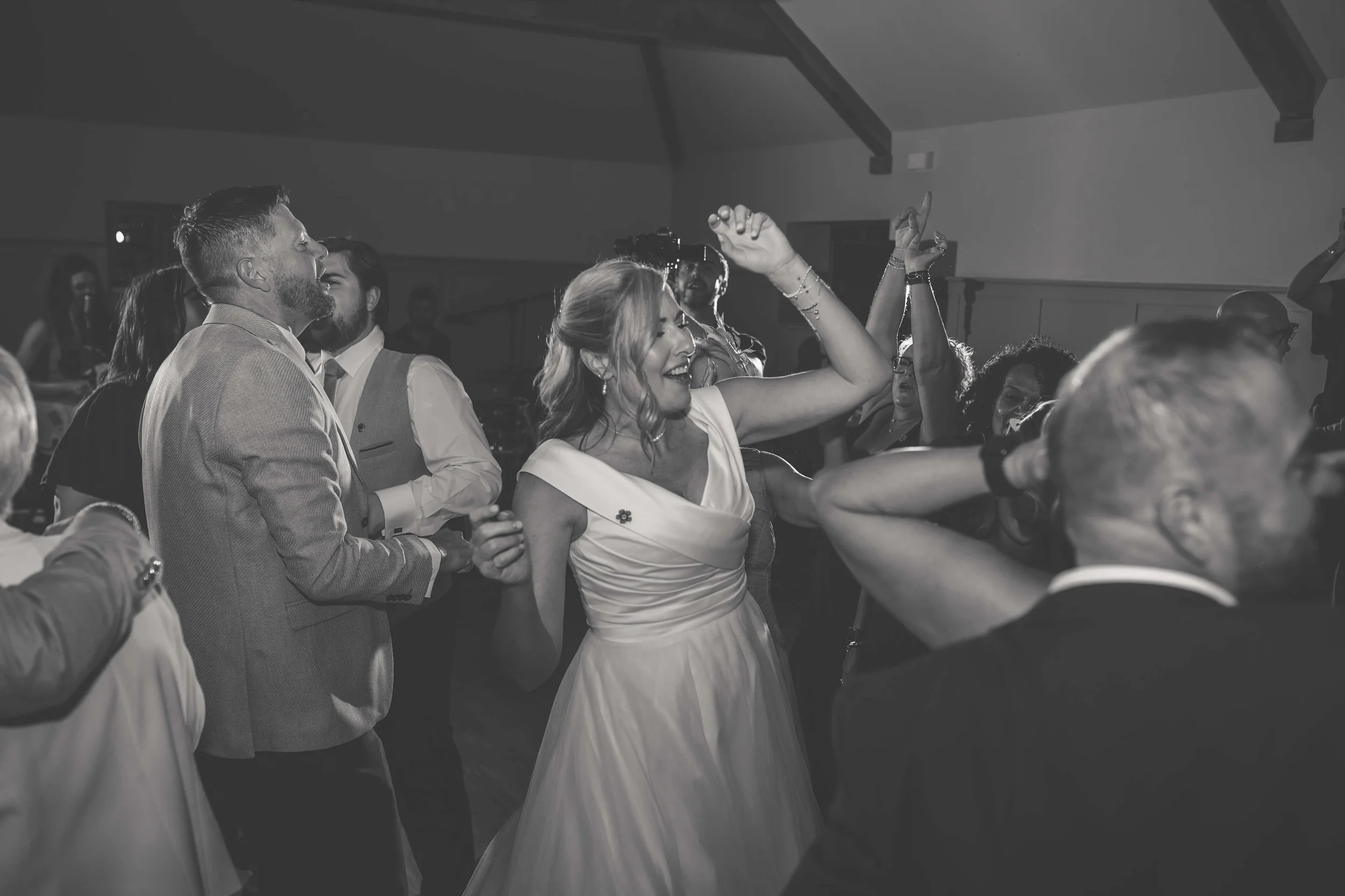 People dancing and celebrating at a wedding reception in a dimly lit indoor venue, with a woman in a white dress smiling and raising her arm.