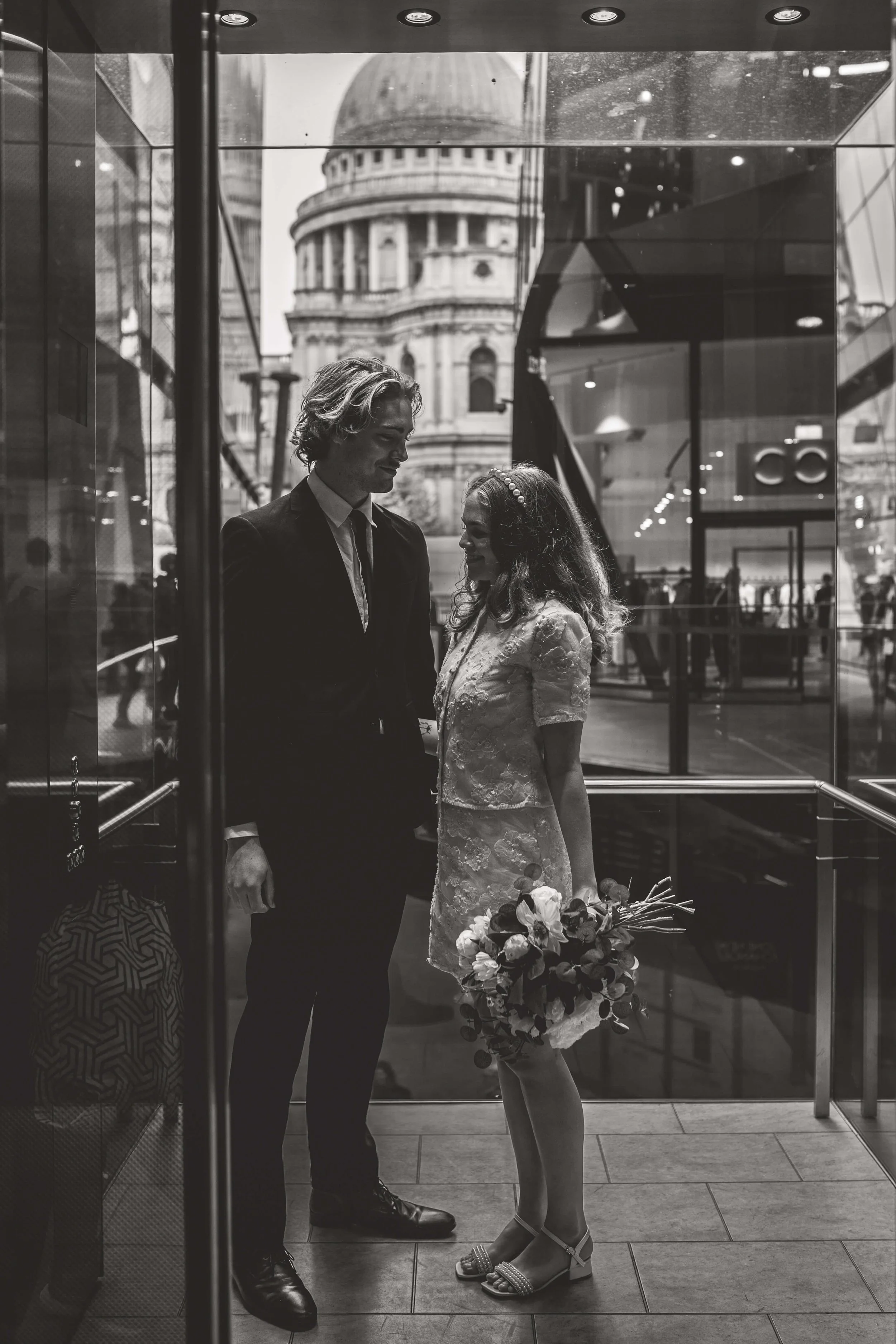 A black and white photo of a couple in an elevator, with a cityscape and historic building visible through the glass behind them. The woman is wearing a dress and holding a bouquet, while the man is in a suit.