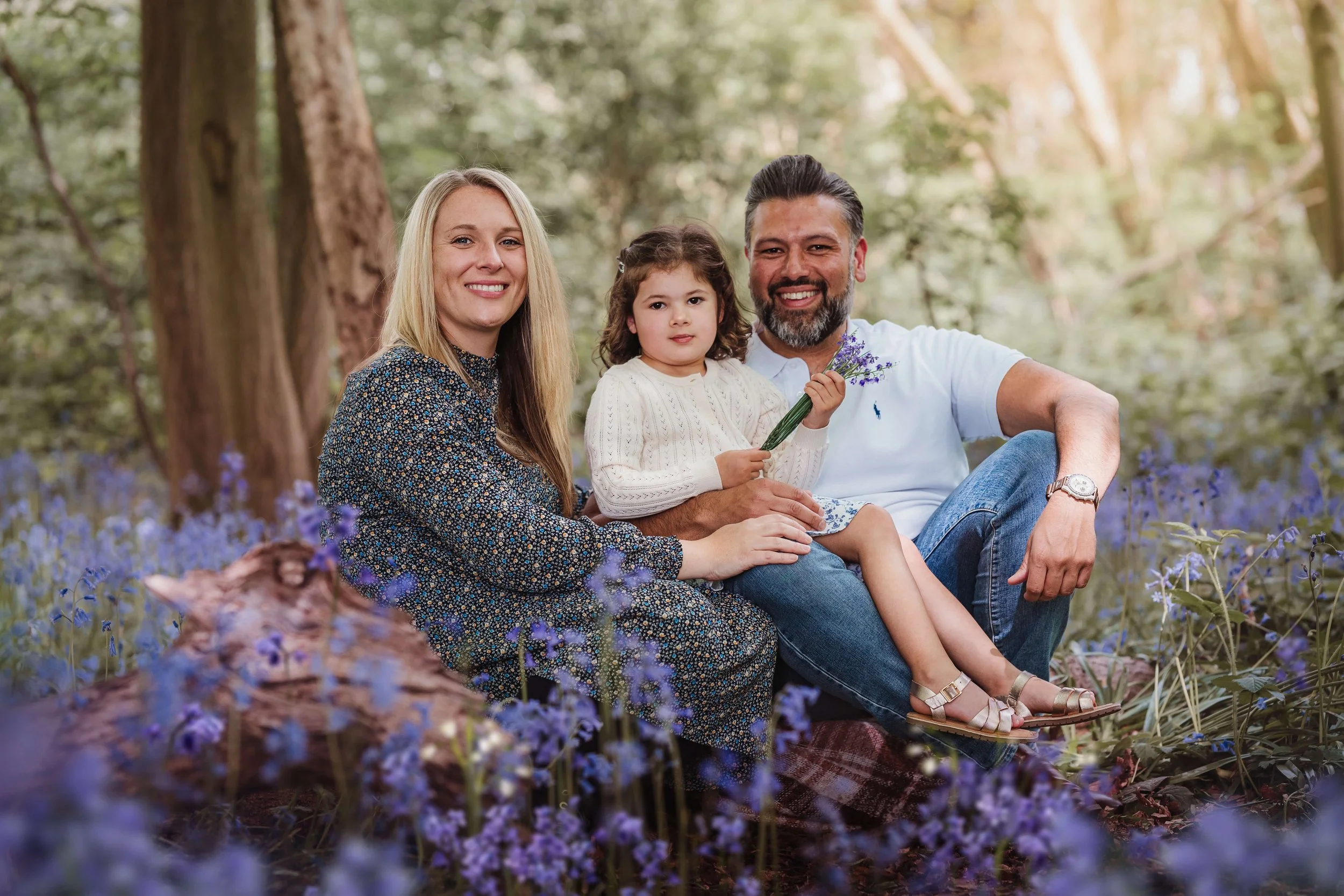 Family of three sitting on a blanket in a bluebell forest, smiling at the camera, with a young girl holding a small bouquet of purple flowers.