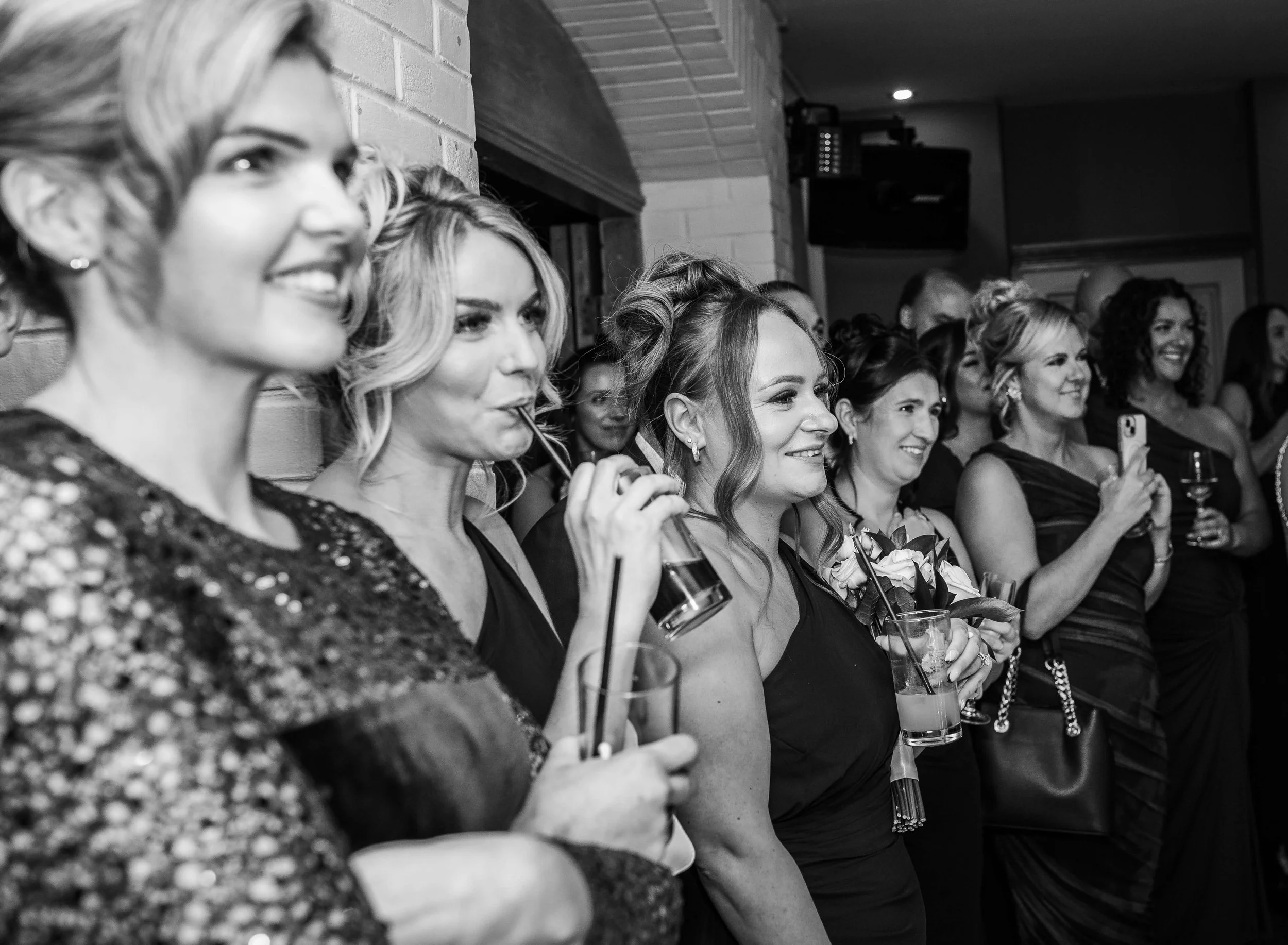 Group of women dressed in elegant black formal attire, standing together at a social event, some holding drinks and one taking a photo with her phone.