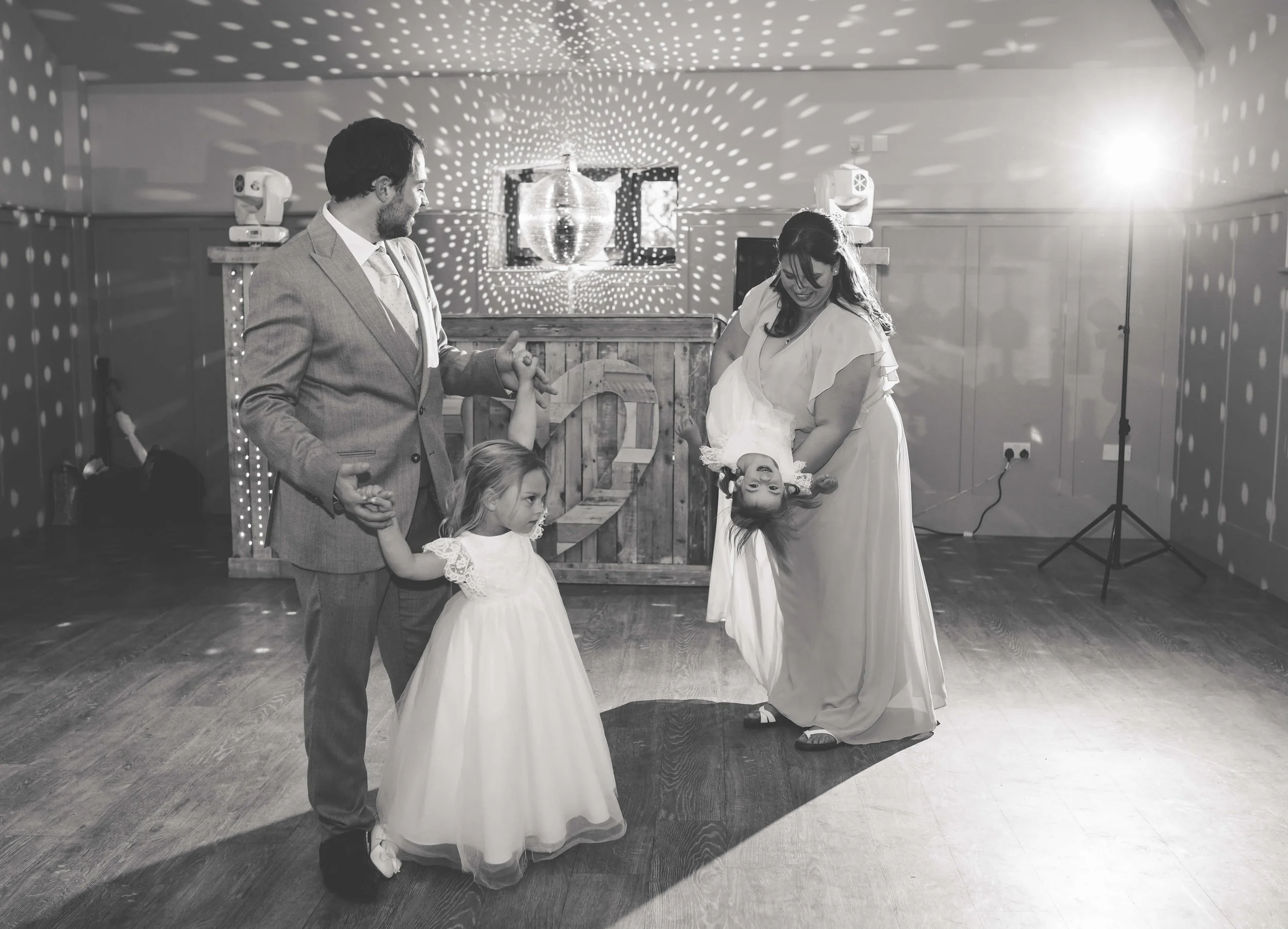 A black-and-white photo of a family dancing at a celebration, with a man, a woman, and two young girls in fancy dresses. The woman is holding one girl upside down while the other girl watches. There are decorative lights and a DJ booth in the backgro