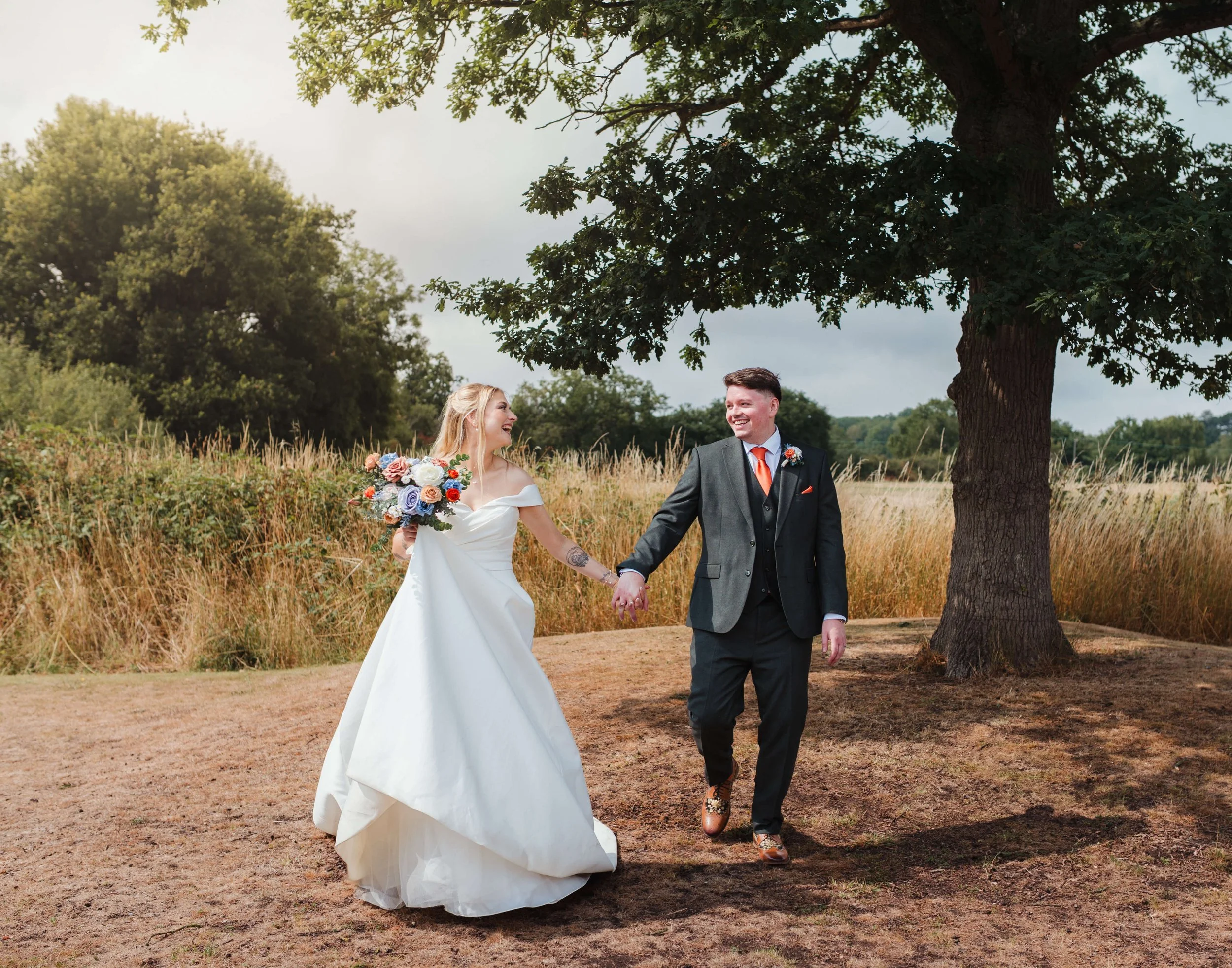 A bride in a white wedding dress holding a colorful bouquet and a groom in a dark suit and orange tie holding hands, walking together outdoors under a large tree in a grassy field.