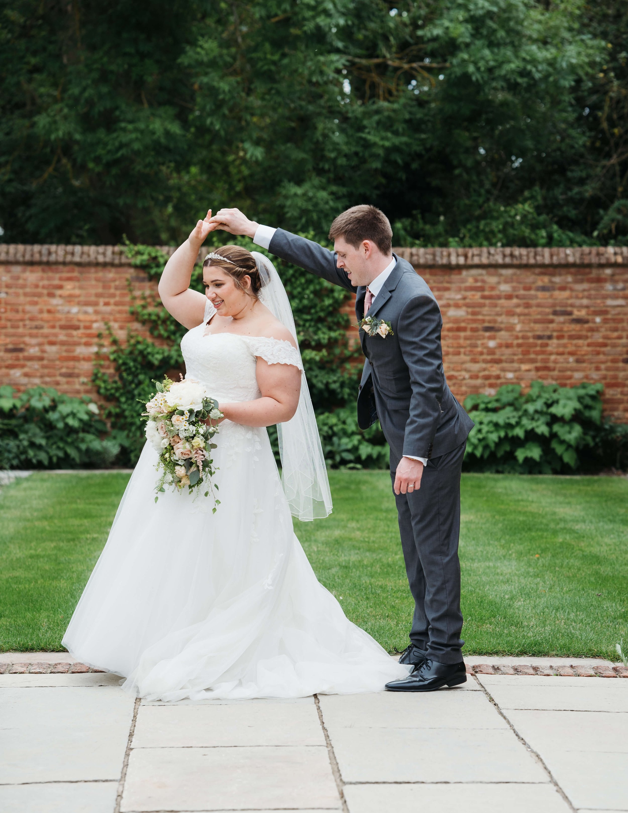 A bride in a white wedding dress and a groom in a gray suit dancing outside on a grass and concrete pathway with a brick wall and green trees in the background.