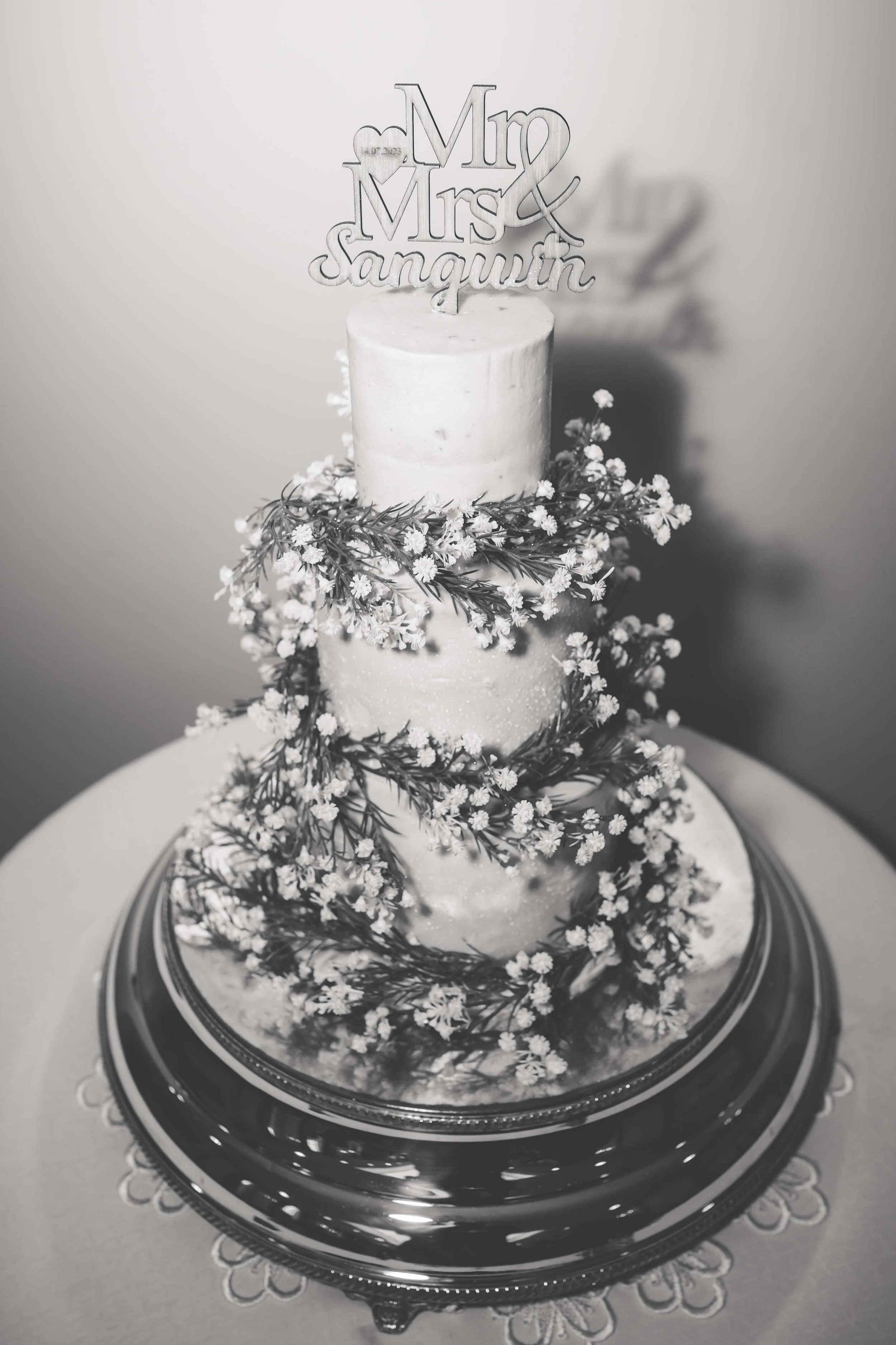 Black and white photo of a three-tier wedding cake decorated with small flowers and greenery, with a topper that says 'Mr & Mrs Sangwin'.