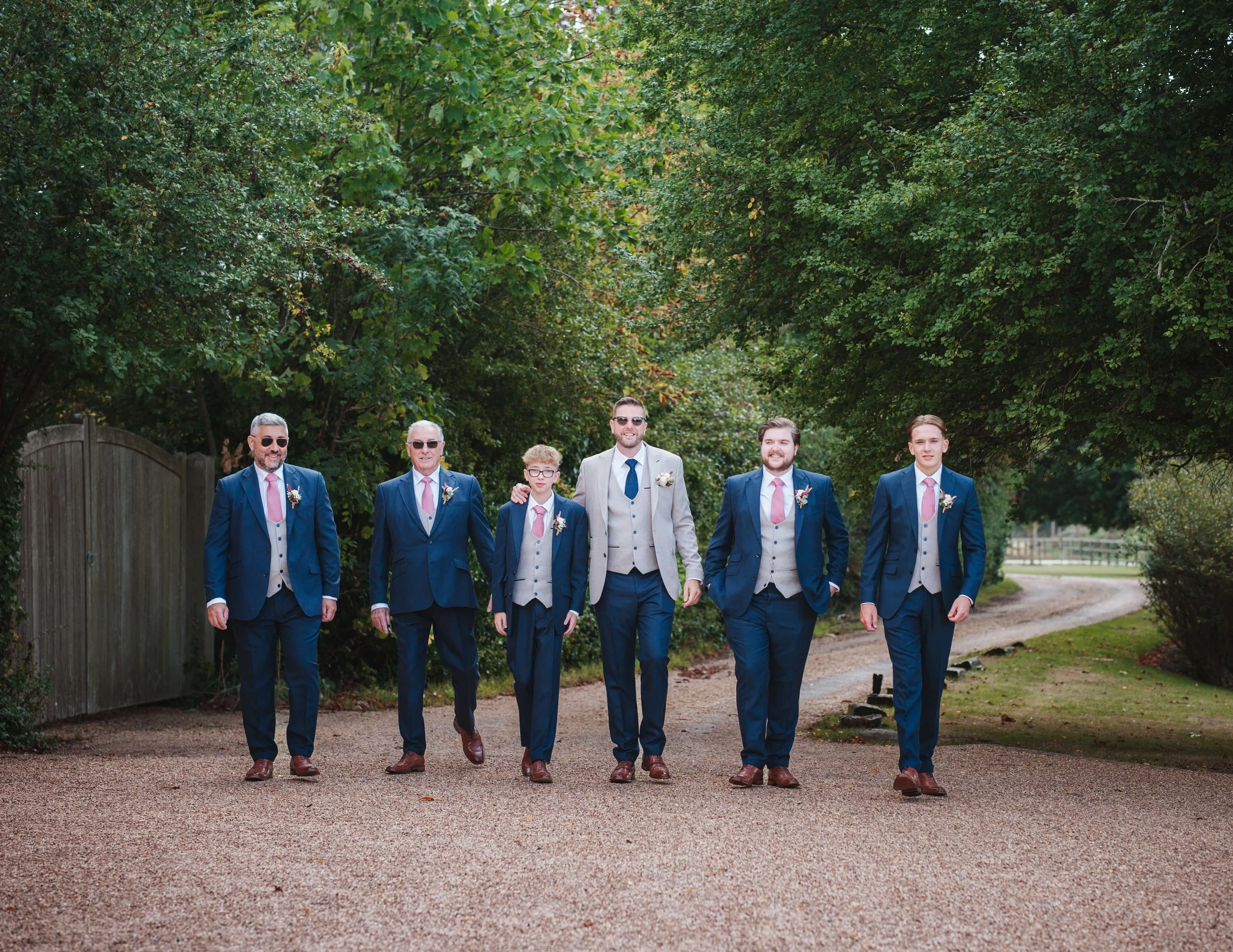 A group of seven men in suits walking outdoors in a lush green park, dressed in matching blue and gray suits, with pink ties and boutonnières, smiling and enjoying the day.