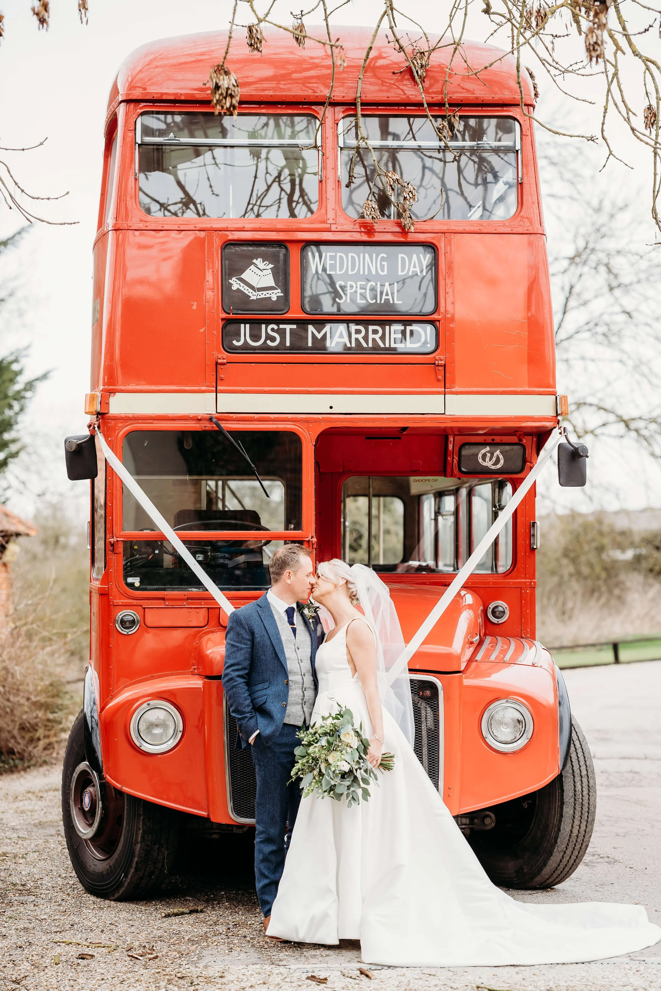 A newlywed couple in wedding attire kissing in front of a vintage red double-decker bus decorated with wedding signs.