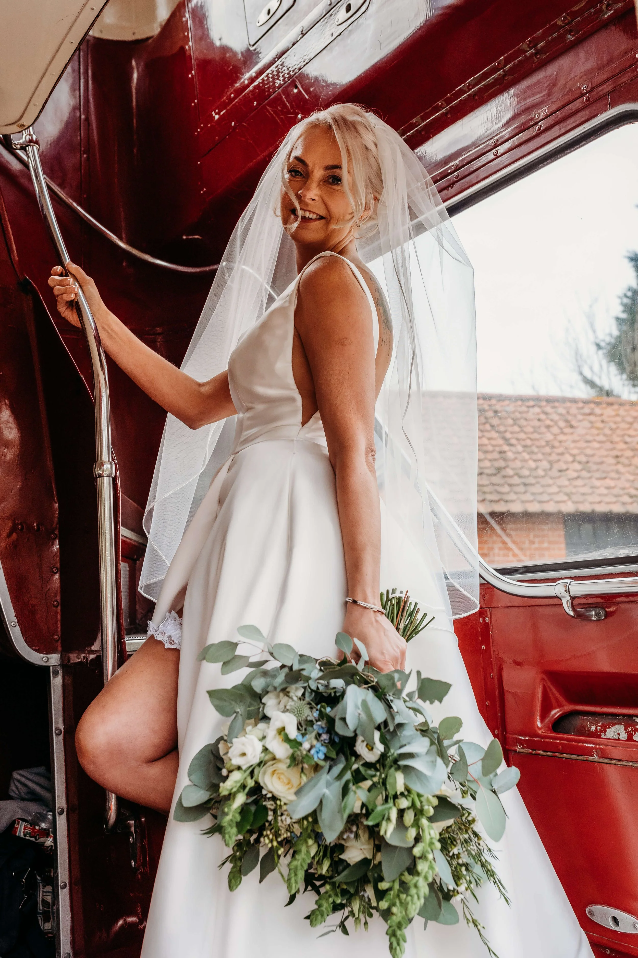 A bride with blonde hair, wearing a white wedding dress and veil, holding a bouquet of white and green flowers, standing inside a vintage red bus.