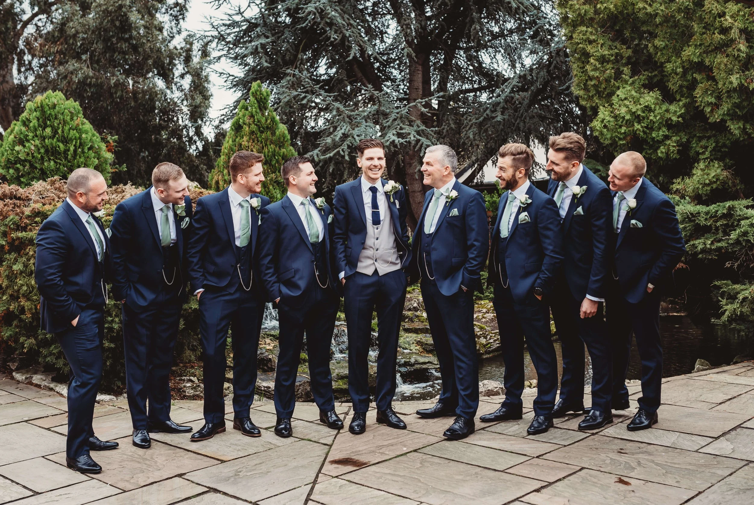 A group of eleven groomsmen dressed in matching navy blue suits, light blue shirts, and mint green ties, standing outdoors on a stone-paved area surrounded by trees and greenery. They are smiling and looking at each other.
