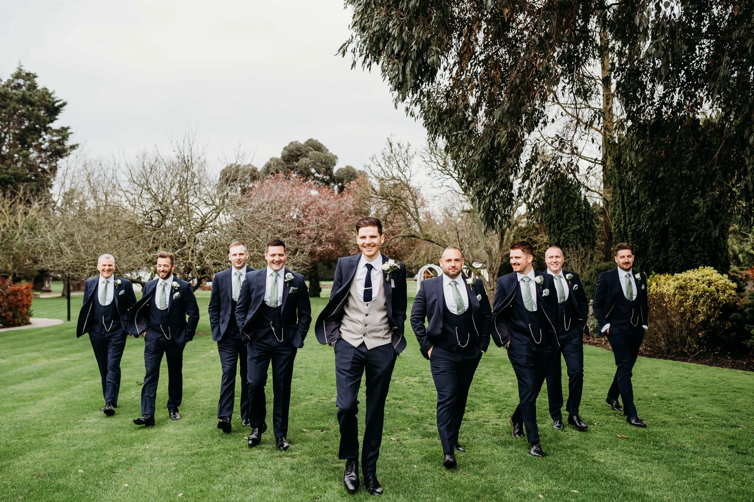 A groom and his groomsmen walking on grass in a park, all dressed in dark suits with light-colored ties, with trees and overcast sky in the background at wedding venue.