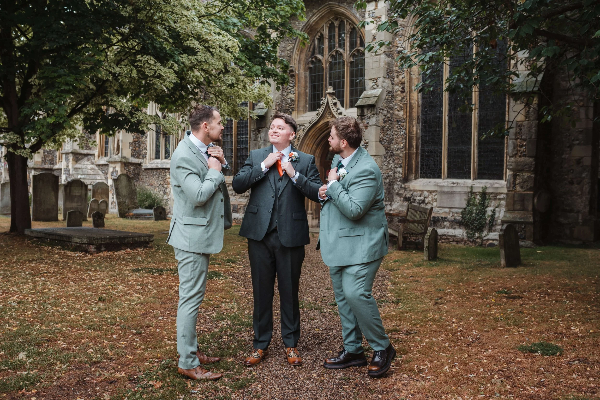 Three men in suits standing outside a historic church, adjusting ties and looking at each other, surrounded by trees and graves.