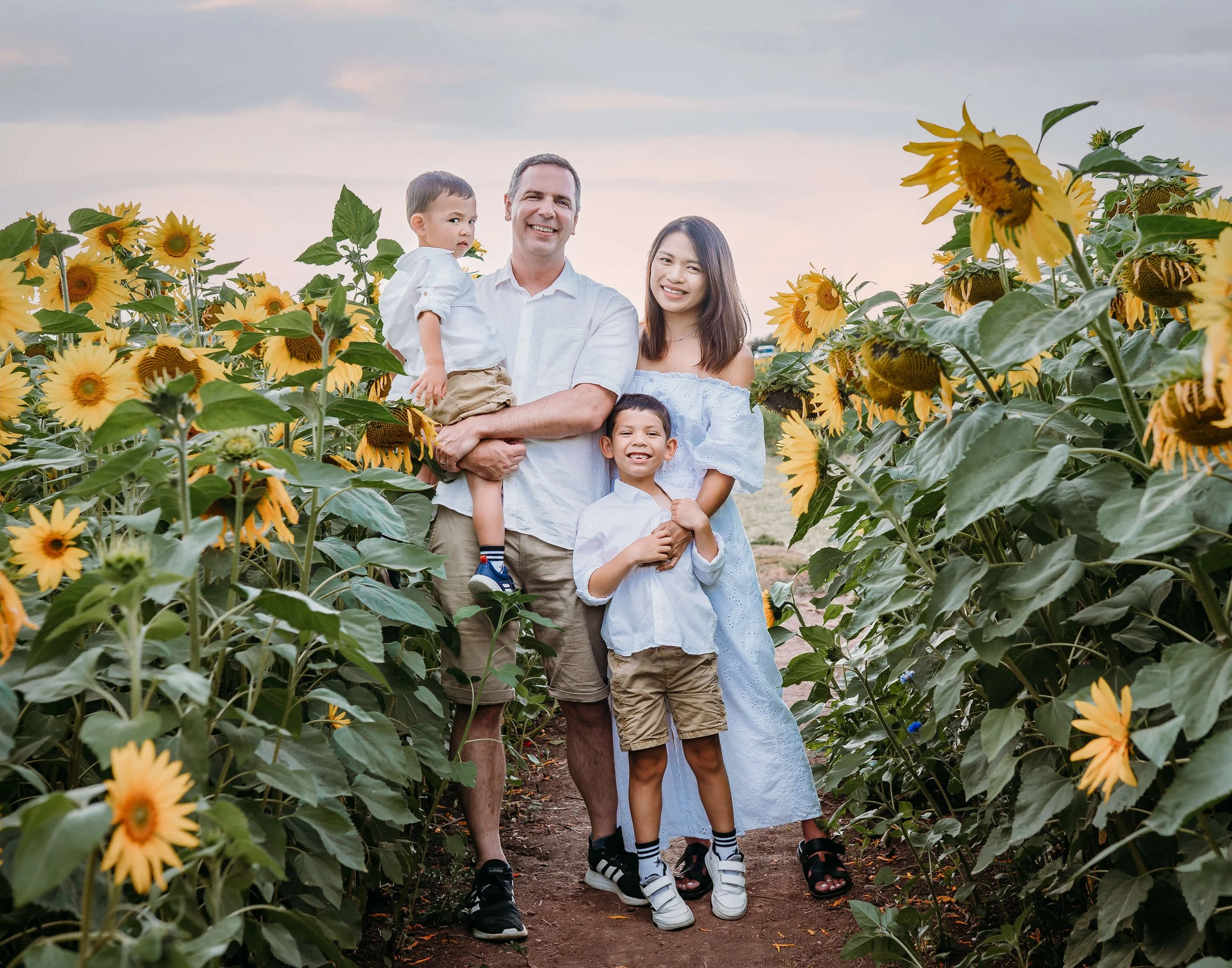 A family of five standing in a sunflower field, smiling at the camera during sunset.