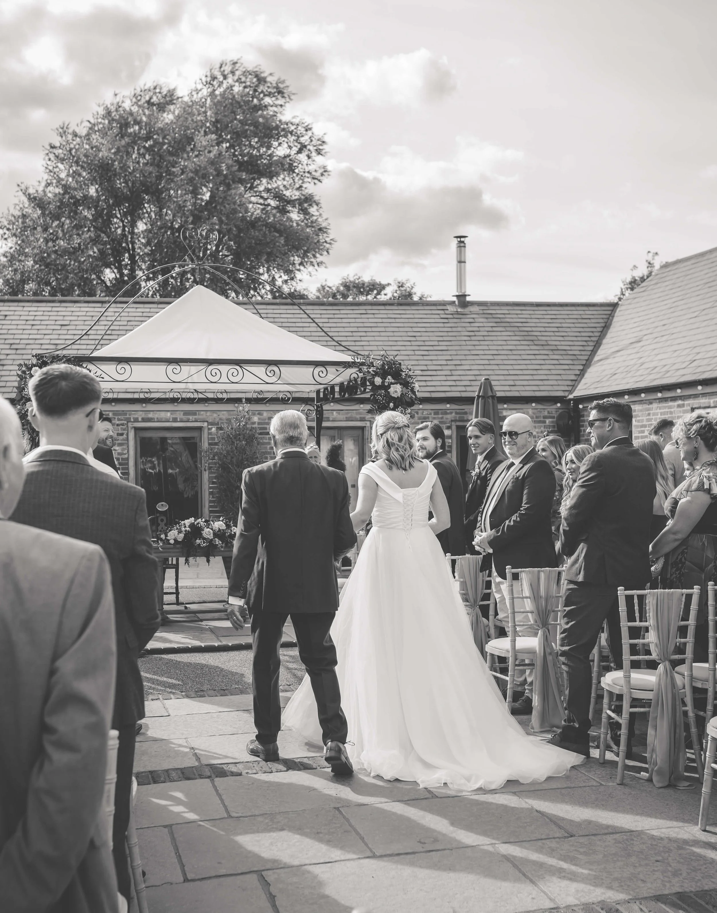 A wedding ceremony outdoors with a bride in a white gown walking with her father, surrounded by guests in formal attire, under a canopy decorated with flowers, on a sunny day.