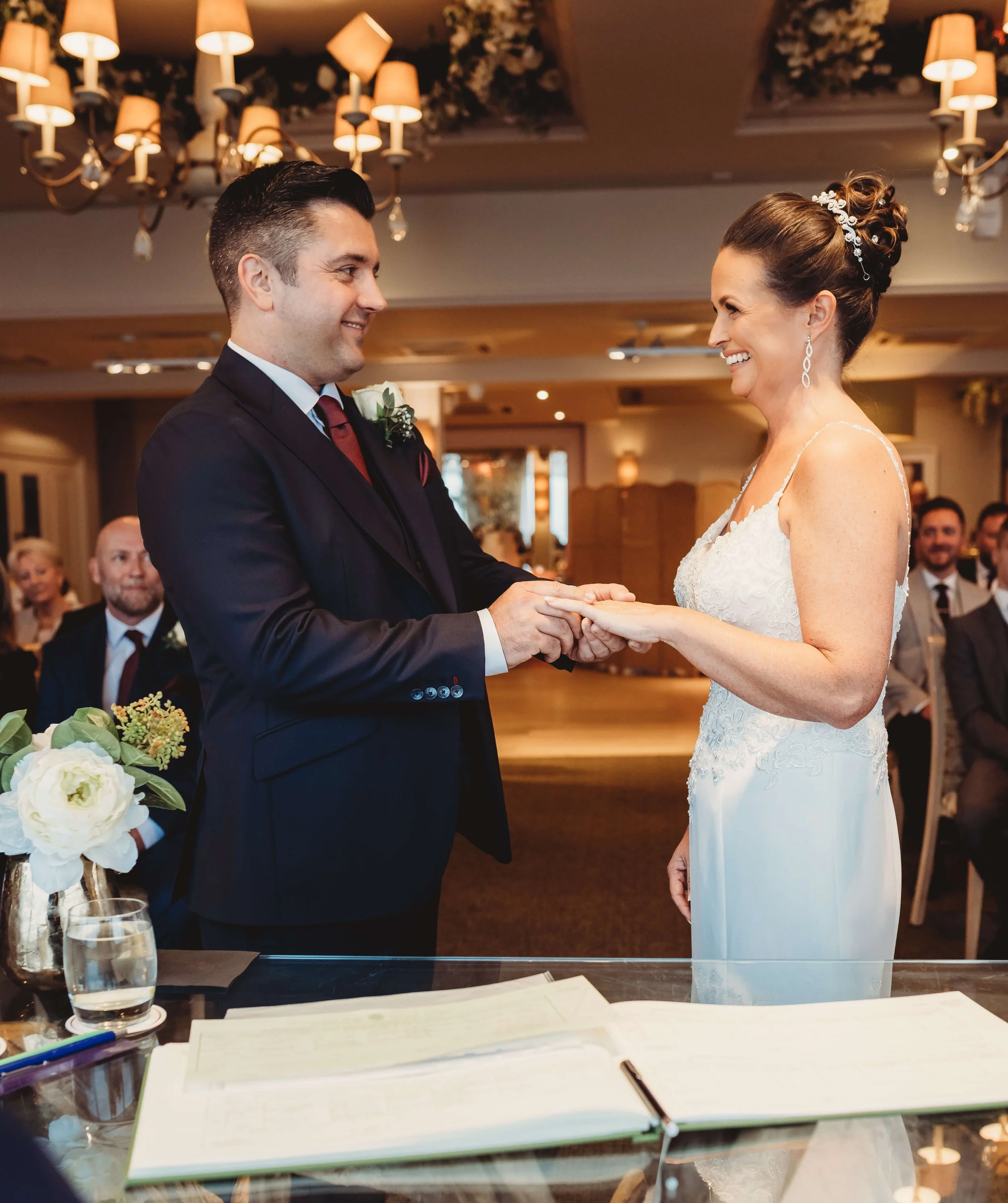 A couple getting married exchanging rings during their wedding ceremony, with guests watching in the background.