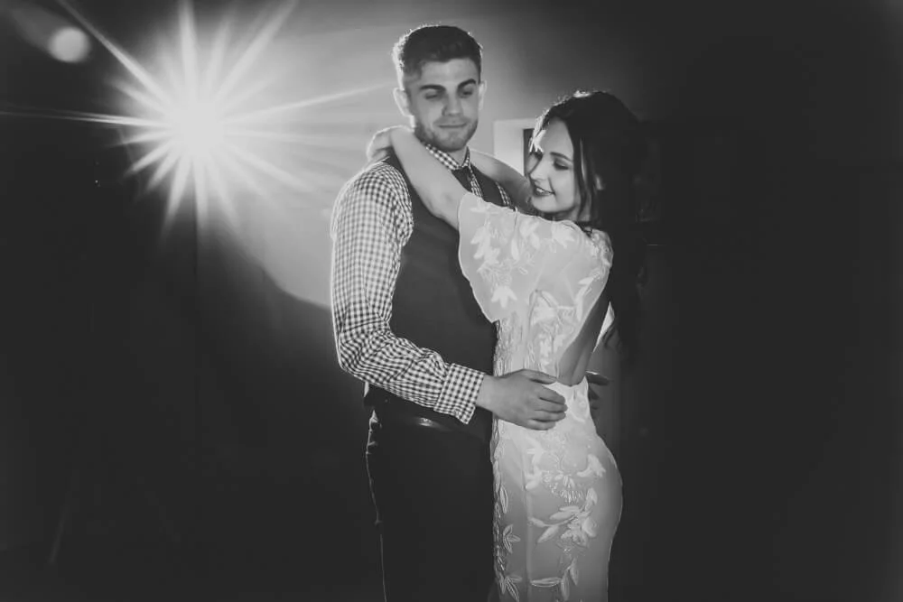 A black and white photo of a couple dancing together, with the woman in a floral dress and the man in a checkered shirt and vest, illuminated by a bright light in the background.