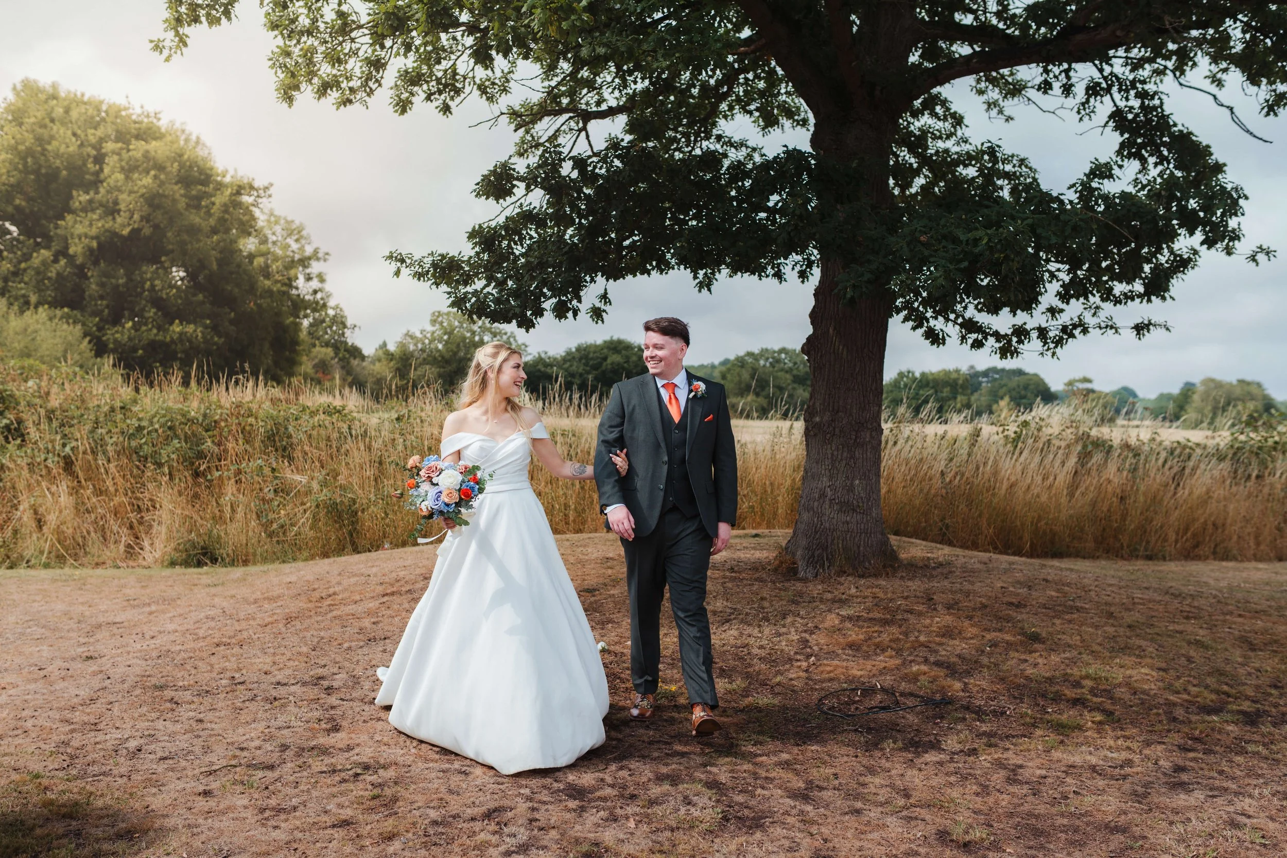 Bride and groom walking hand in hand under a large tree outdoors, smiling at each other during their wedding celebration.