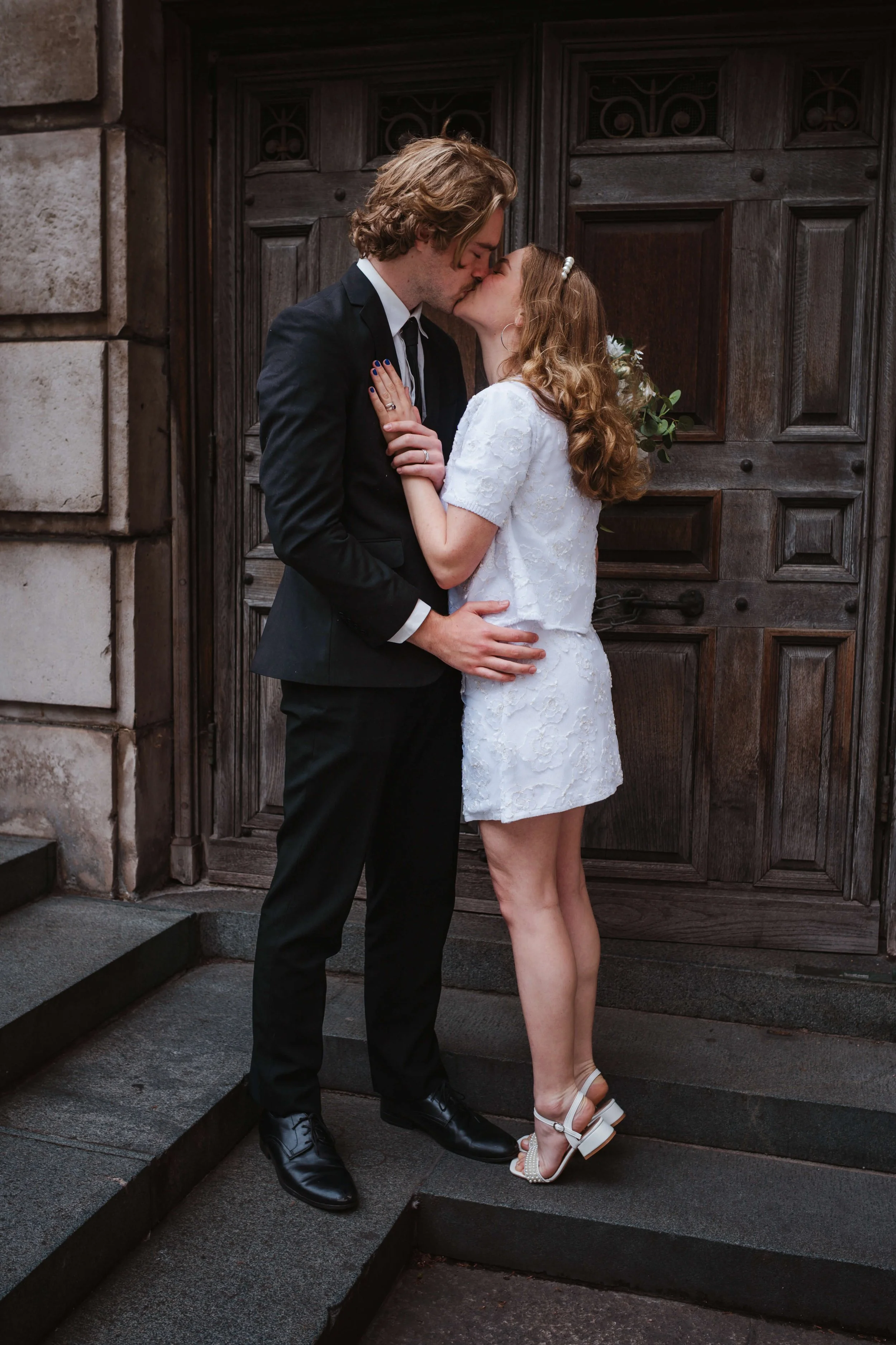 A couple dressed in formal attire sharing a kiss on the steps outside a building with large wooden doors.