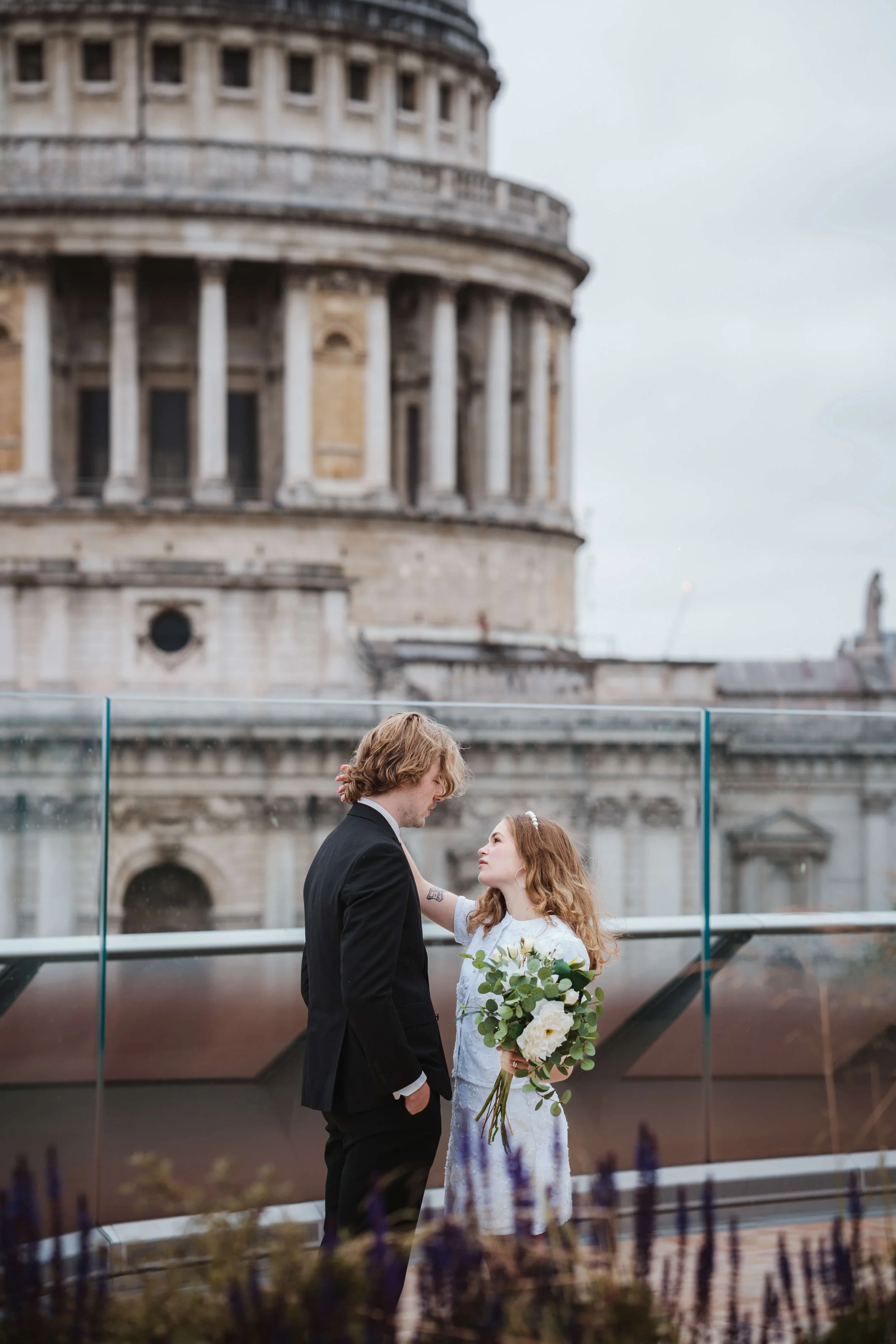 A couple on a rooftop overlooking a historic building, with the woman holding a bouquet of flowers and the man dressed in a suit.