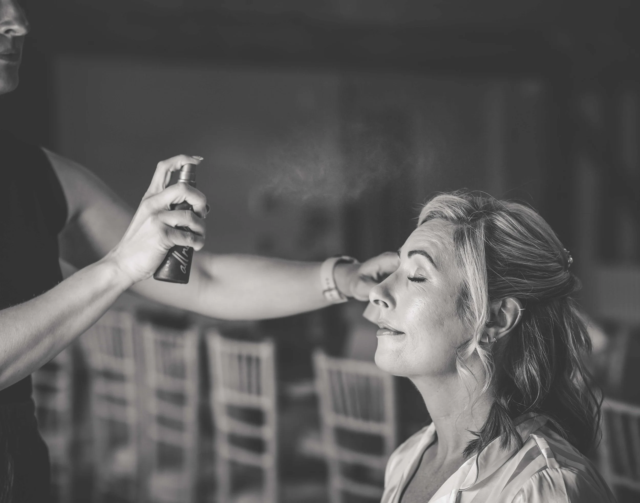 A woman getting her makeup done, having eyes closed, while a makeup artist sprays mist on her face.