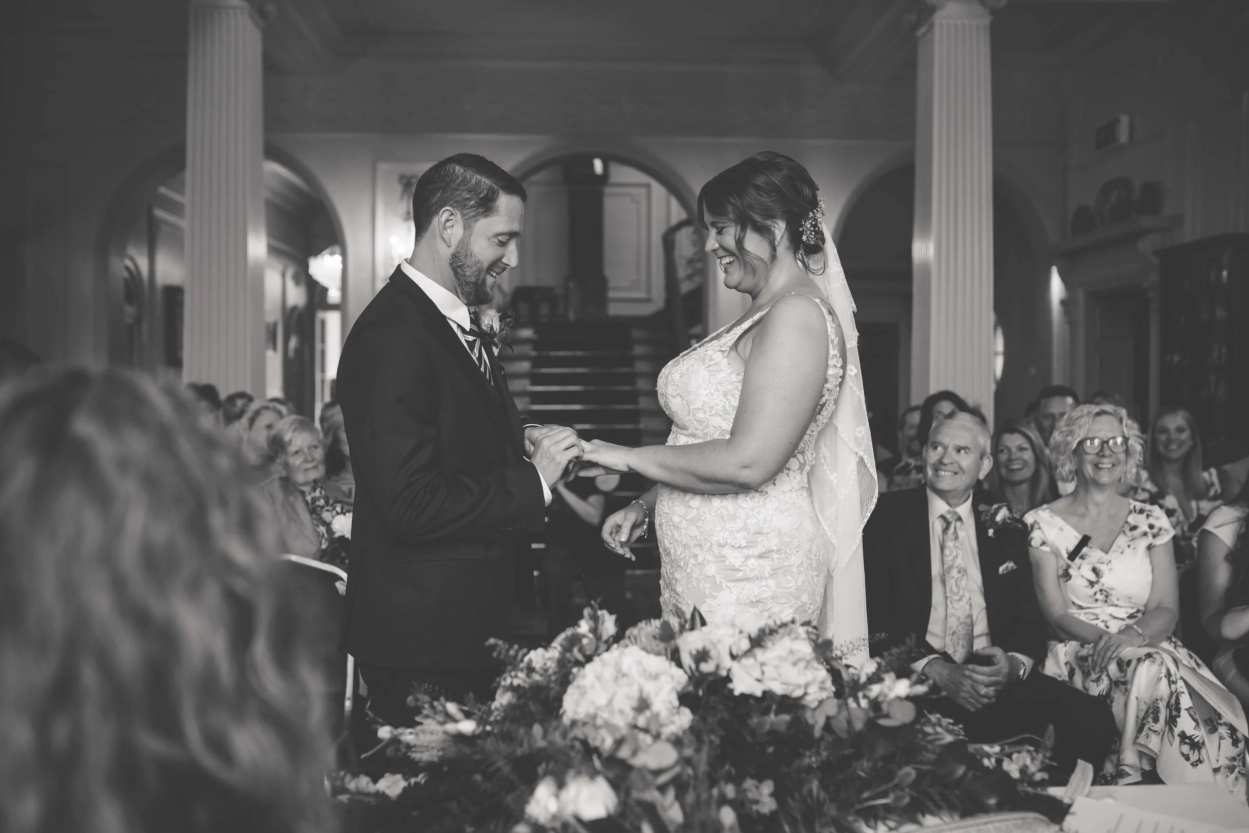 A black-and-white photo of a wedding ceremony with the bride and groom exchanging rings. The bride is wearing a lace wedding dress with a veil, and the groom is in a suit. Guests are seated and smiling in the background.