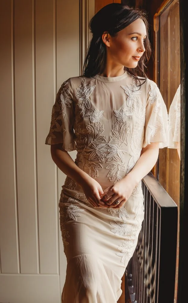 A woman in a beige dress with embroidered floral and leaf patterns, standing by a window, looking outside, inside a house.